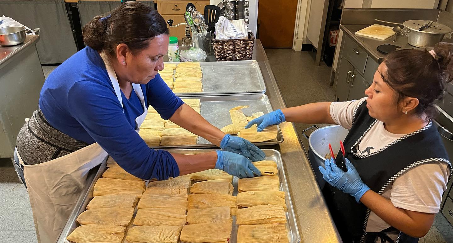 Two women working together in a kitchen, preparing food on a counter with trays of dough, wearing gloves.