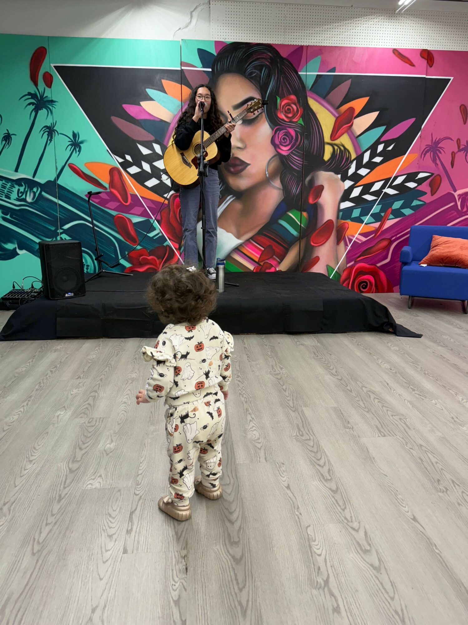 Child standing on light-colored wooden floor in front of colorful mural, holding a guitar, with a black bag nearby.