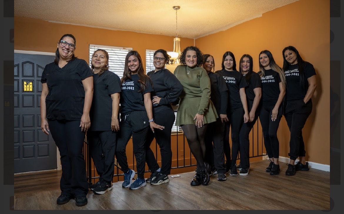 Group of nine women standing indoors, smiling, with orange walls and wooden floor, some wearing matching black shirts.