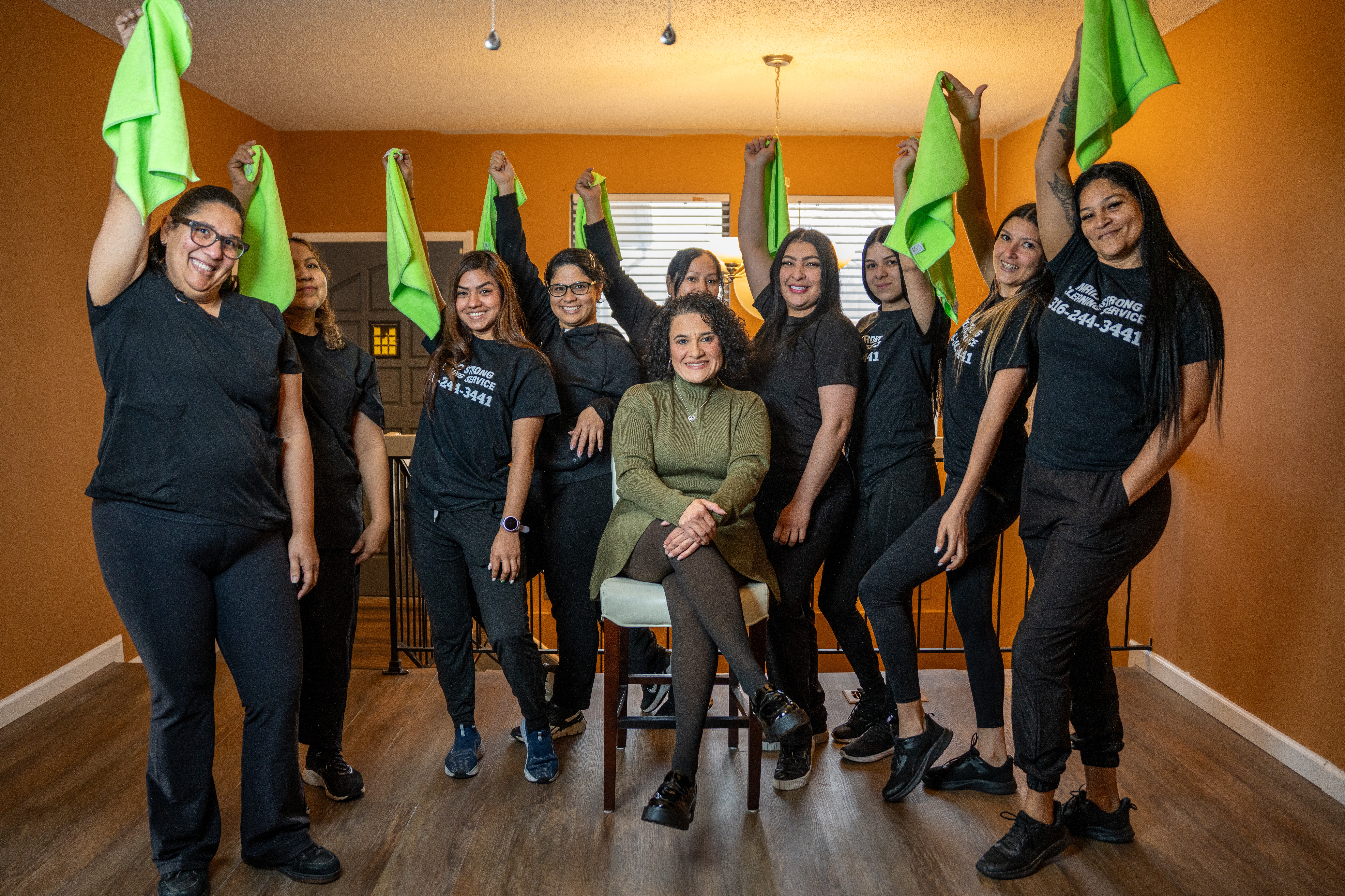 Group of women in black shirts holding green cloths, some smiling, one seated in a chair, in a room with orange walls.