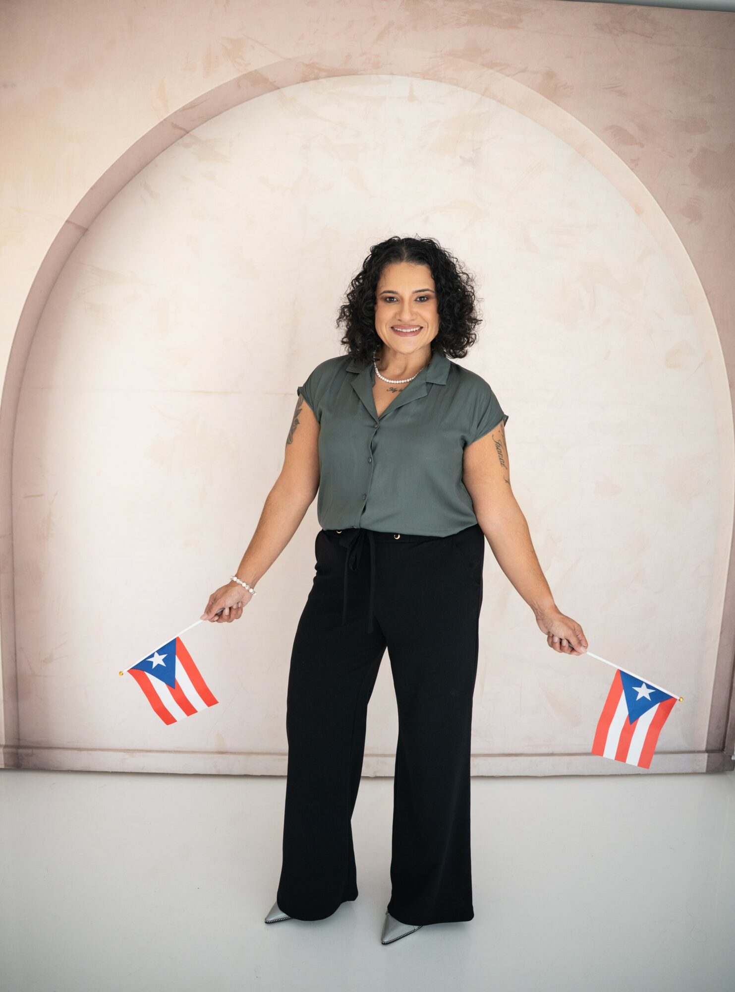 Woman with curly hair smiling, holding Puerto Rican flags, standing in front of a circular backdrop.