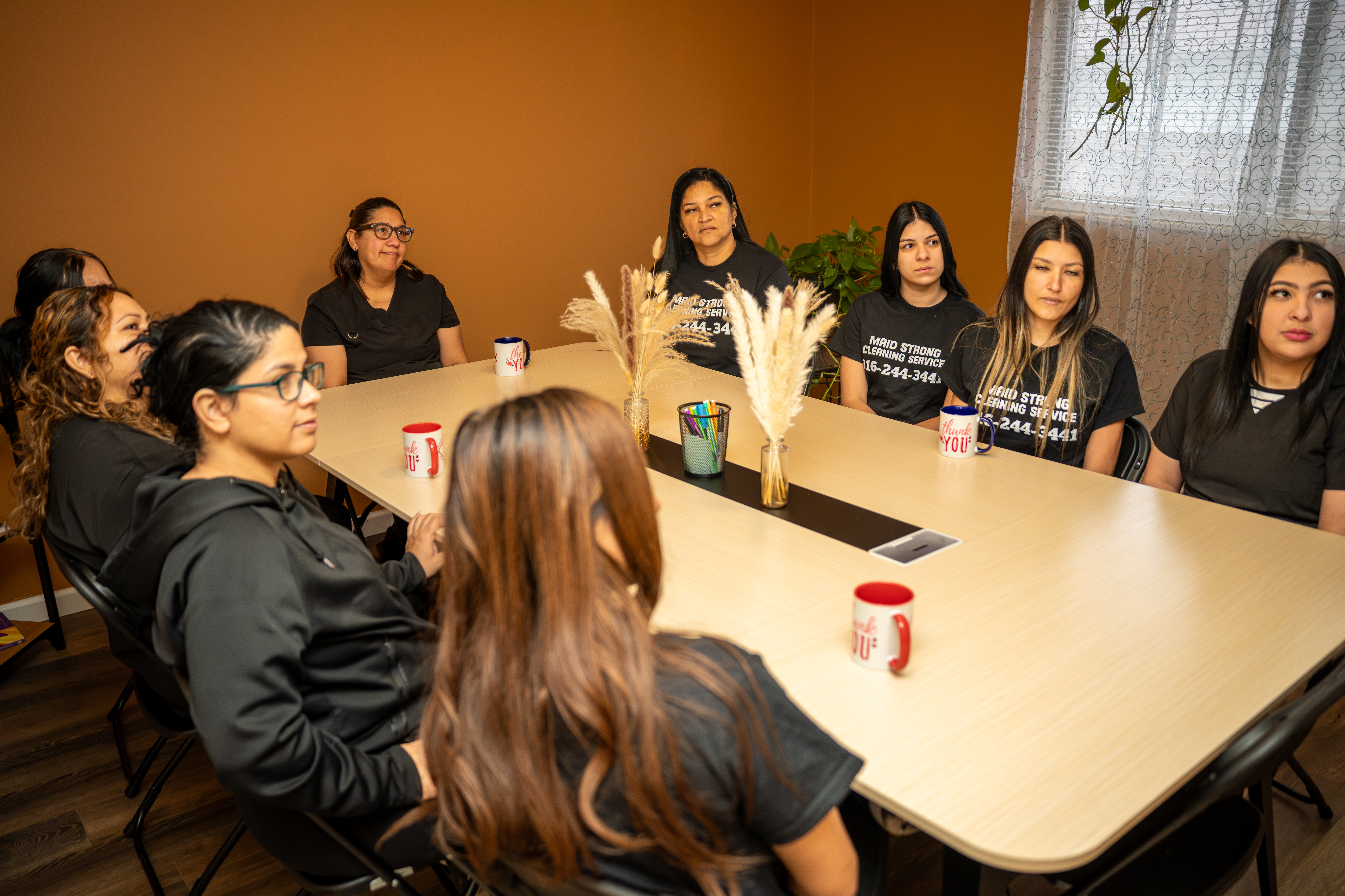 Group of women sitting around a rectangular table in a room with brown walls and a window with curtains.
