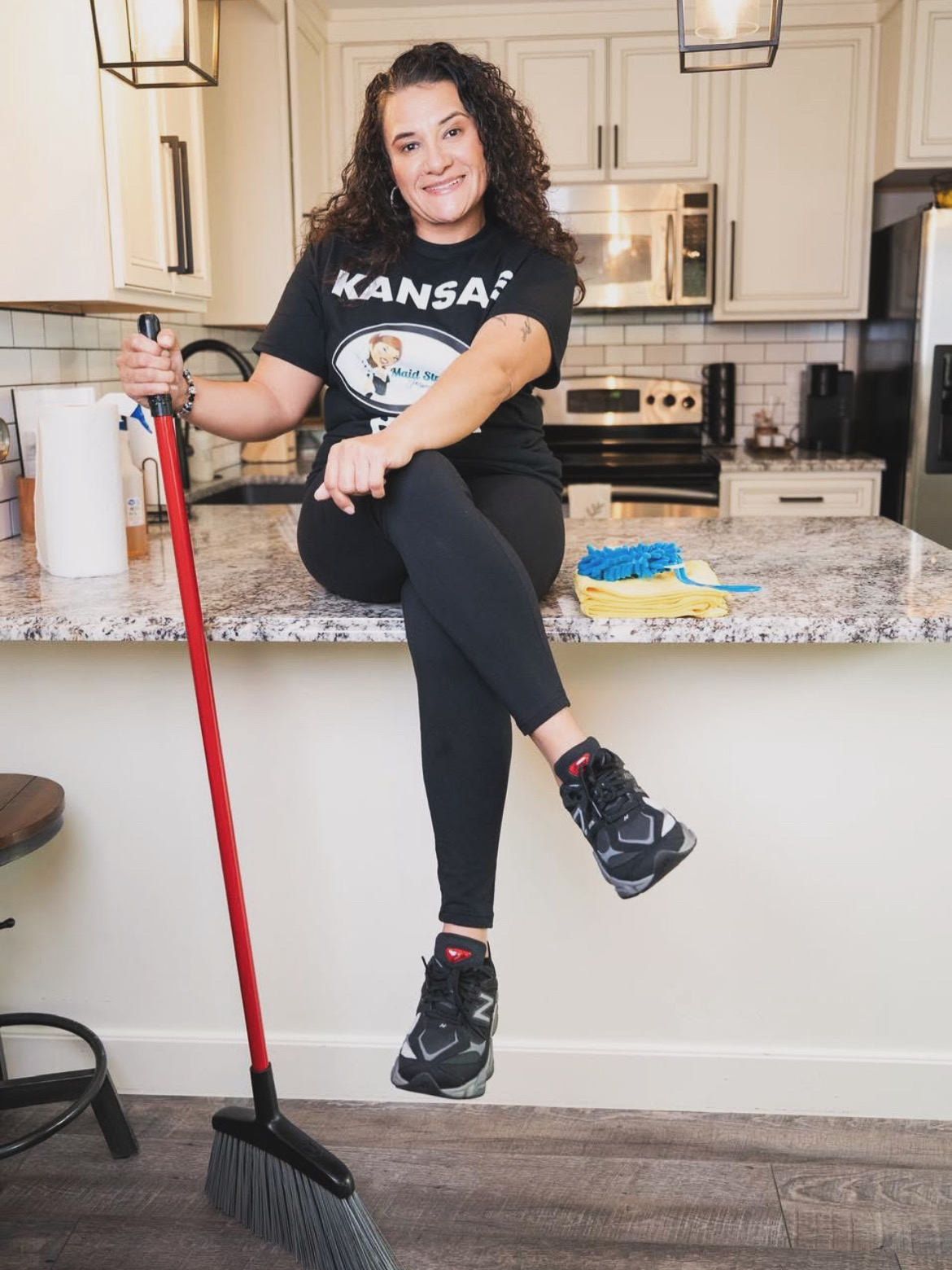 Woman sitting on kitchen counter holding a broom, smiling, with kitchen appliances in background.