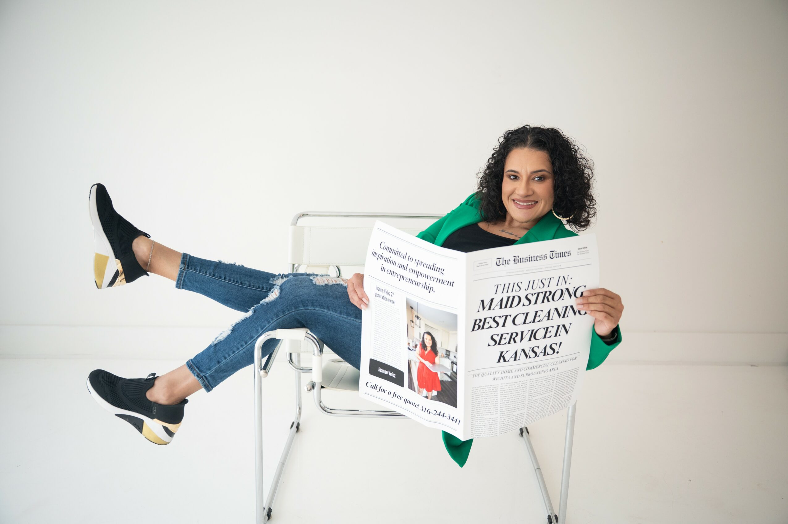 Woman sitting on a chair, reading a newspaper, with legs extended and crossed, smiling, against a plain background.