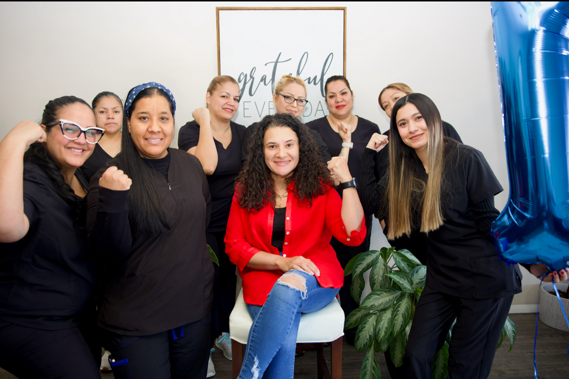Group of nine women smiling, some with fists raised, in a room with a framed sign and a large blue balloon, celebrating.