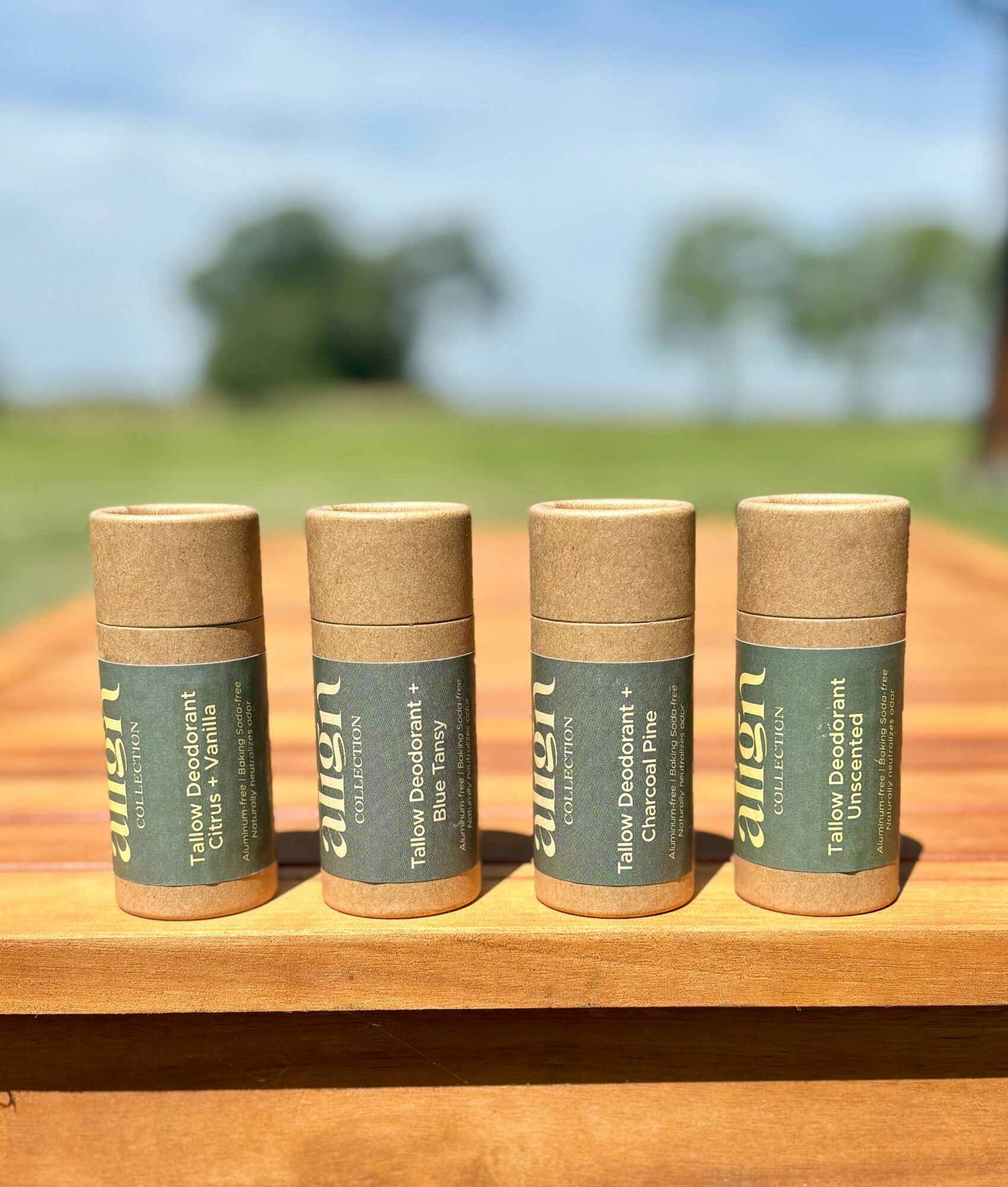 Four cylindrical containers with green labels are placed on a wooden surface outdoors, with trees and a blue sky in the background.