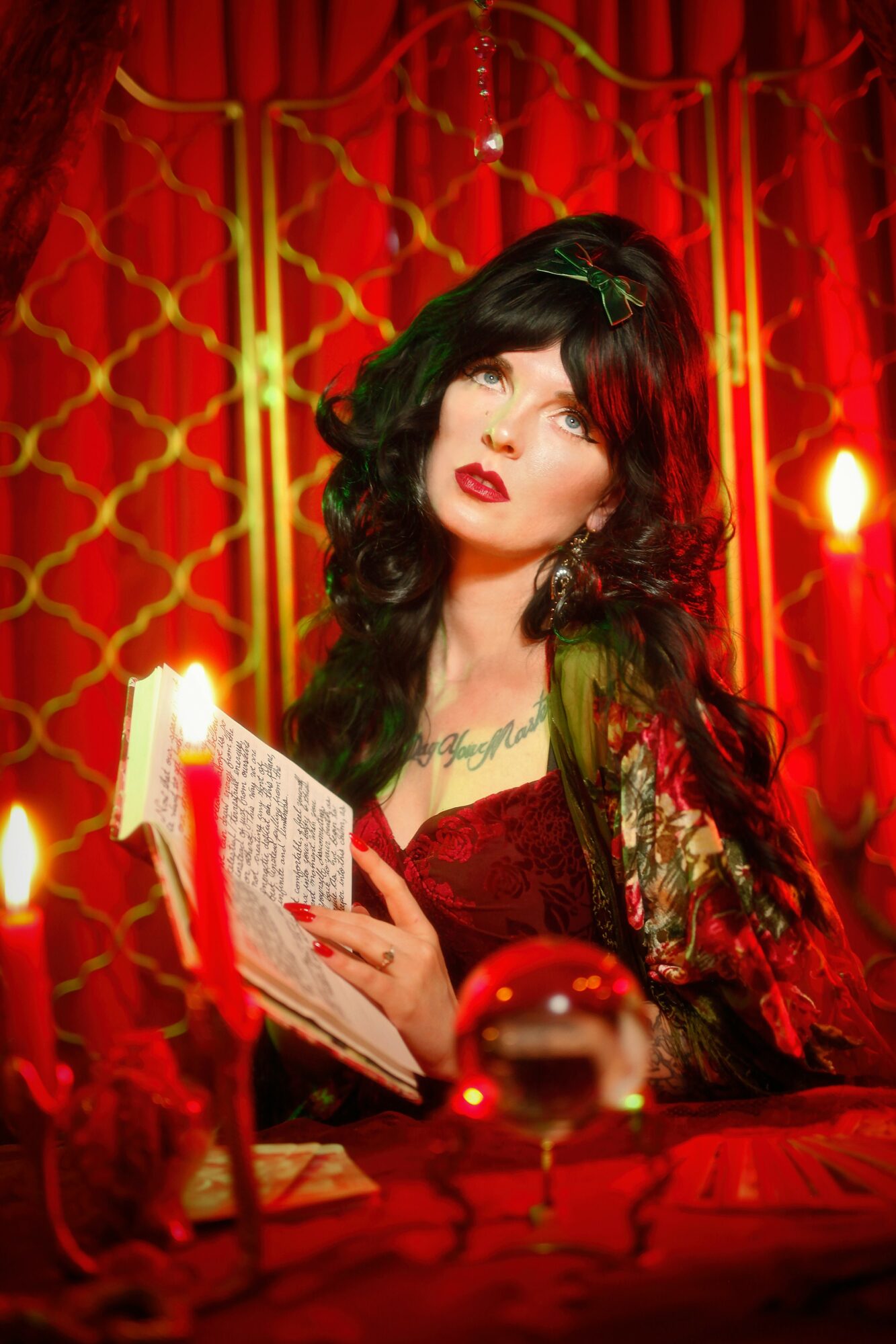 Woman with dark hair and red lipstick reading a book in front of a red curtain, surrounded by candles and decorative objects.