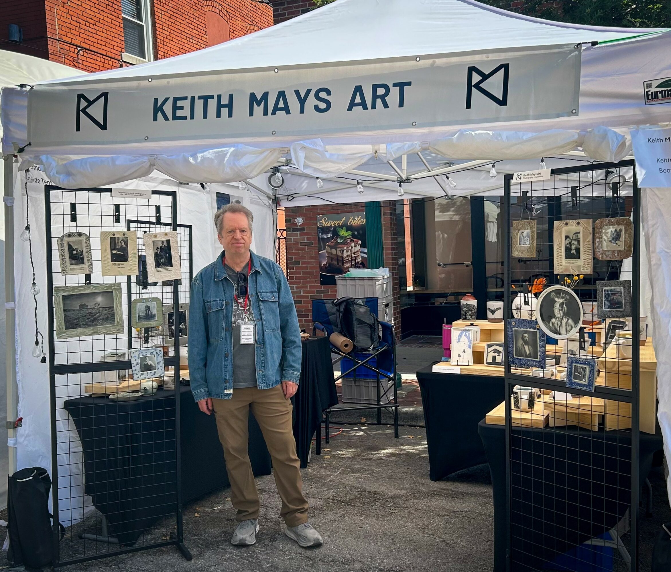 Man standing in front of art display at Keith Mays Art booth, outdoor event, tents, artwork on display, brick building background.