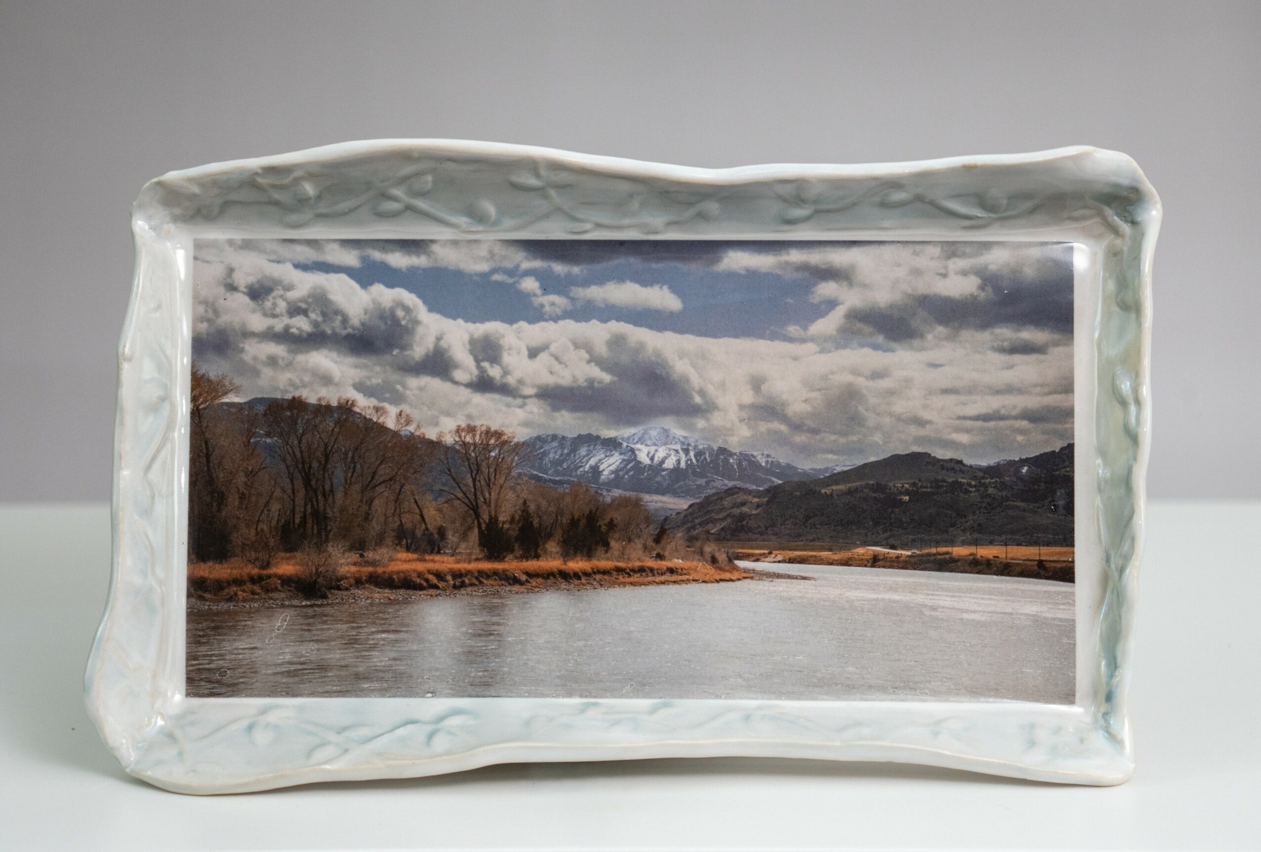 Landscape with river, trees, mountains, and cloudy sky, in a decorative ceramic frame.