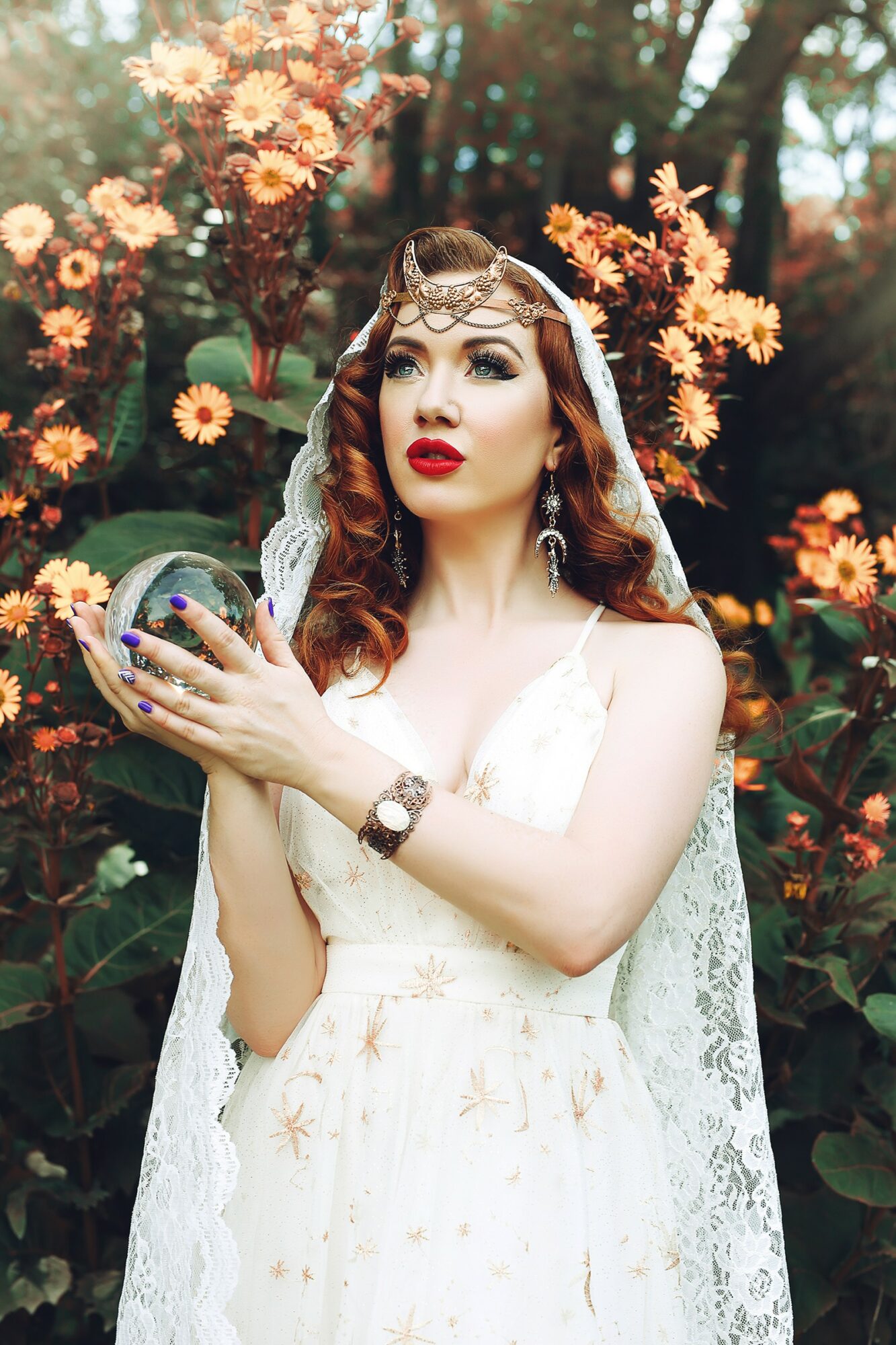 Woman in white dress with lace veil holding a mirror, surrounded by orange and pink flowers, outdoors.