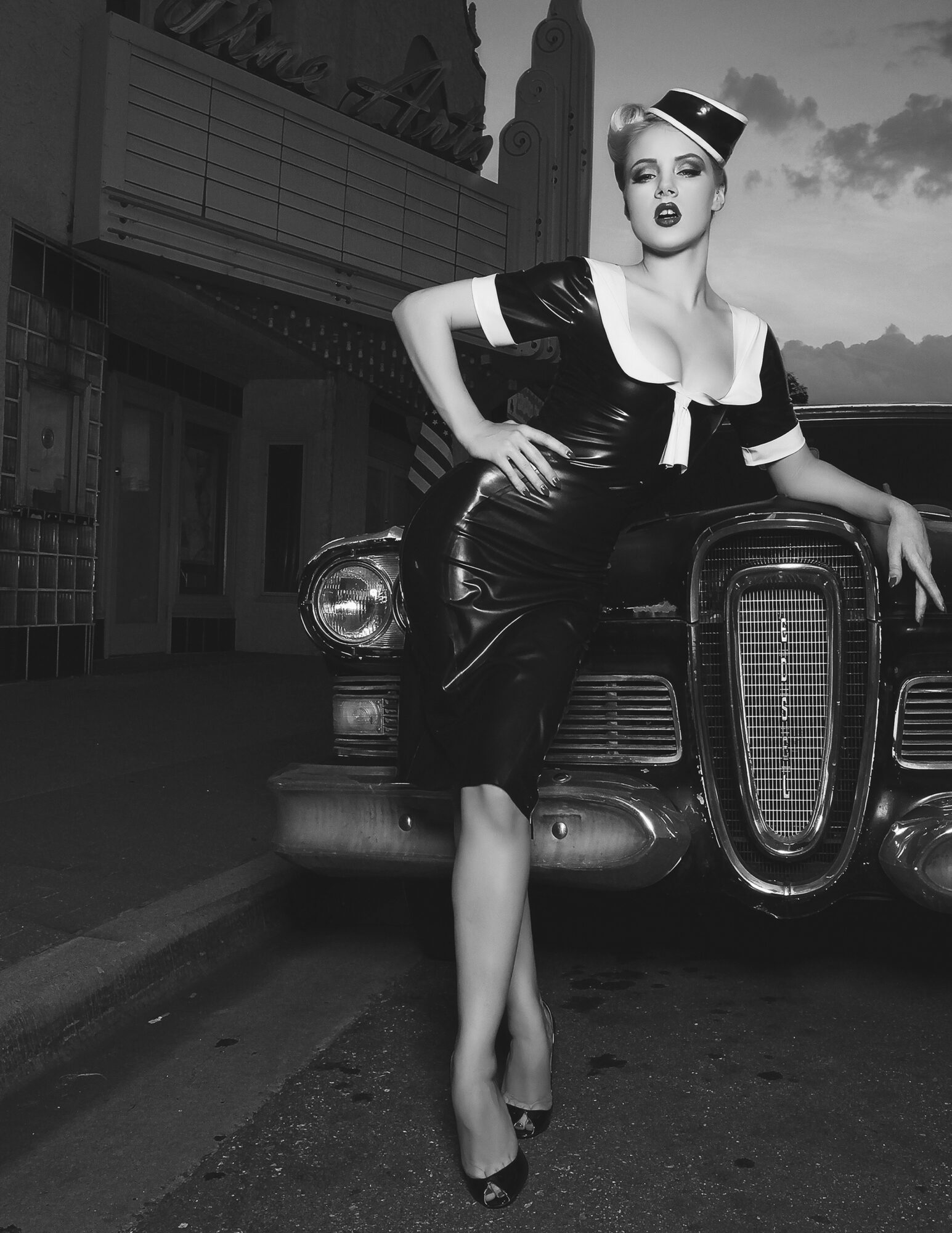 Woman in vintage outfit leaning on a classic car, cityscape background, black and white photo.