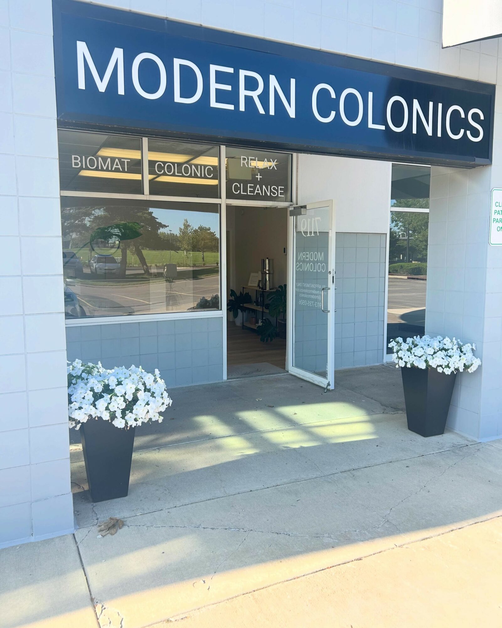 Clinic entrance with open door, two large flower pots with white flowers, and a sign reading 'MODERN COLONICS'.