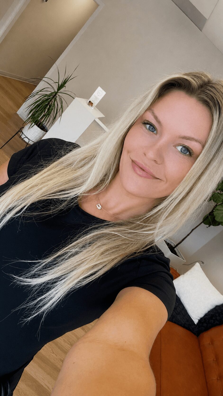 Young woman with long blonde hair and blue eyes smiling in a modern room with plants and furniture