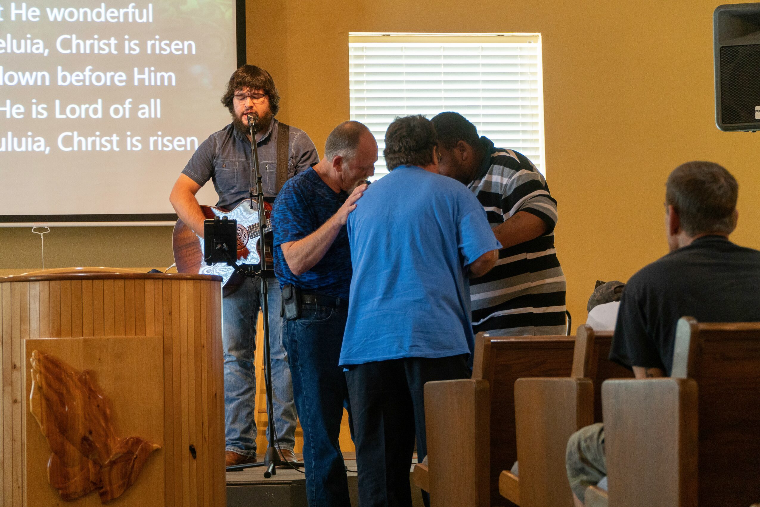 Group of people gathered in a room with a man on stage, a screen displaying lyrics, and others standing and sitting.