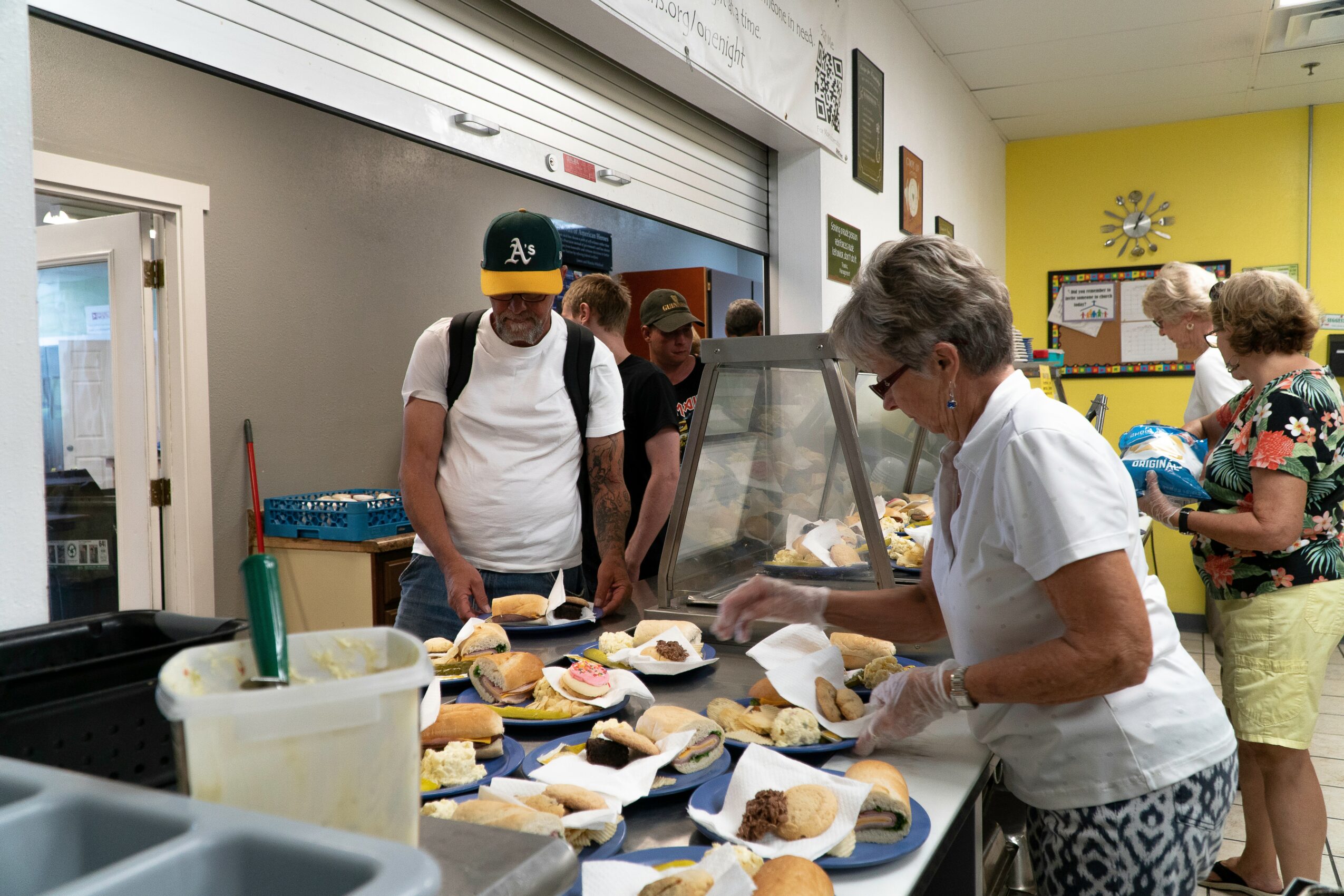 People gather around a table with sandwiches and food, serving and preparing meals in a room with yellow walls.