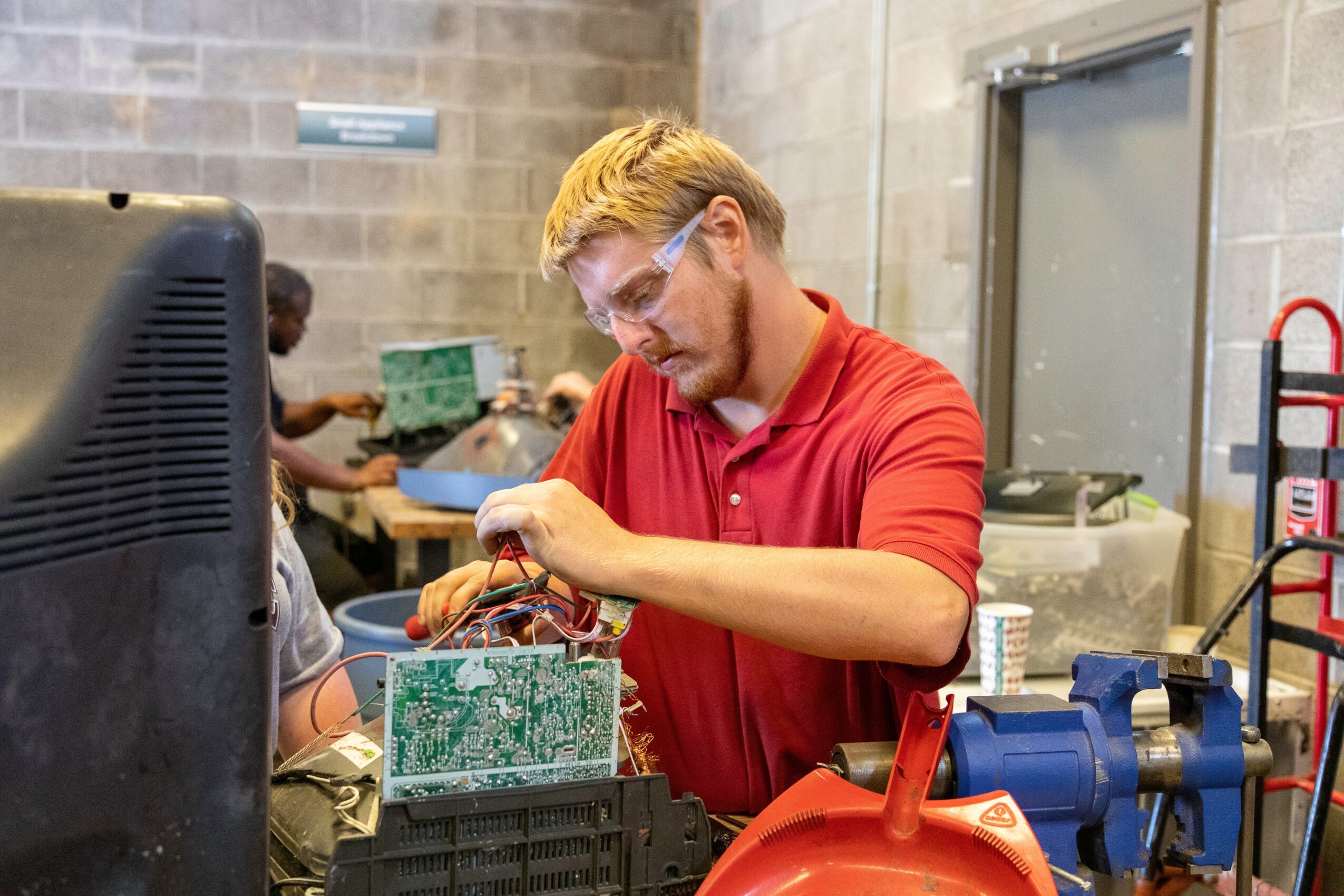 Young man with safety glasses working on electronic components at a workbench in a workshop.