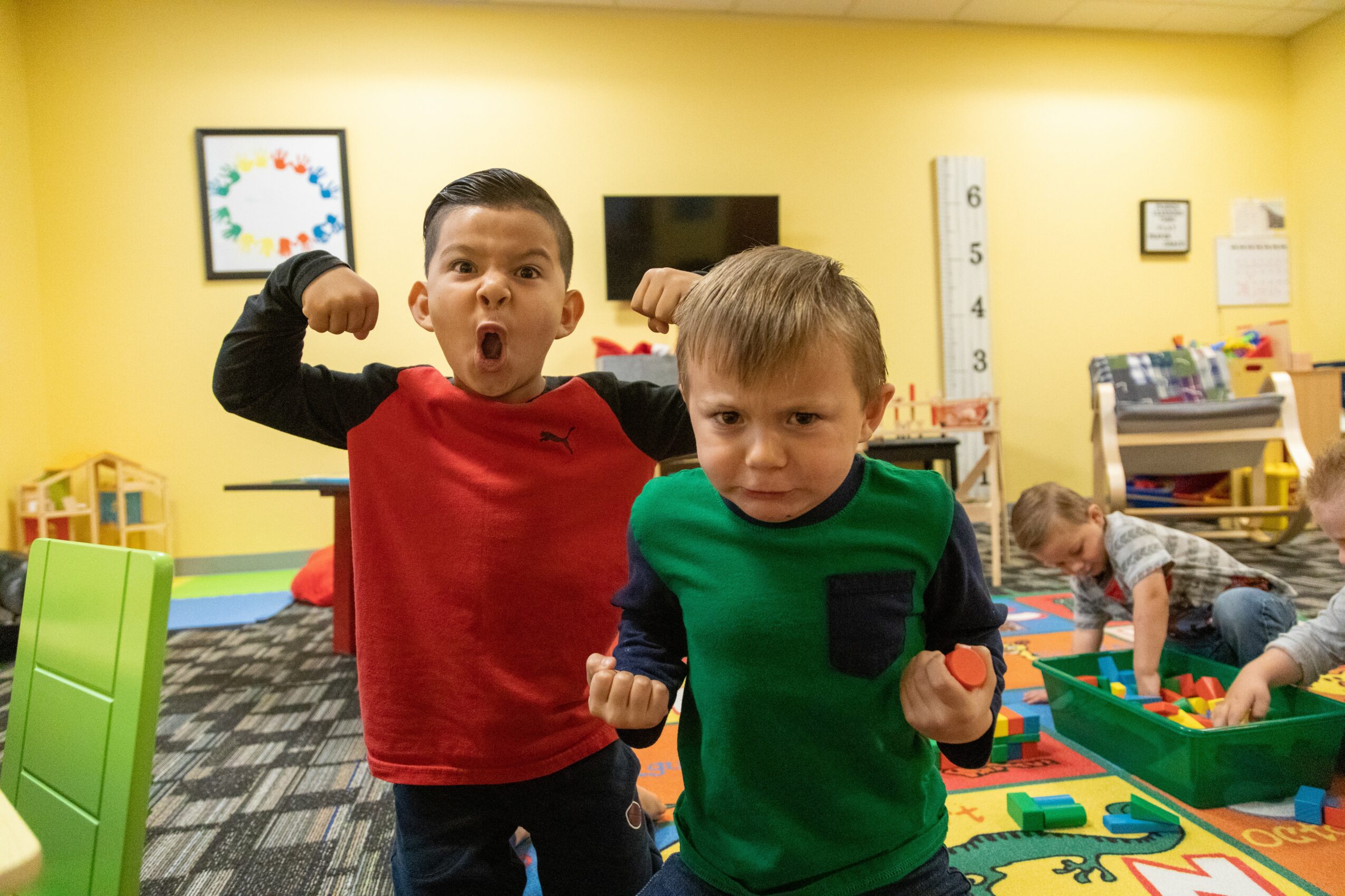Two boys in a colorful classroom, one flexing muscles and the other with fists raised, with children playing in the background.