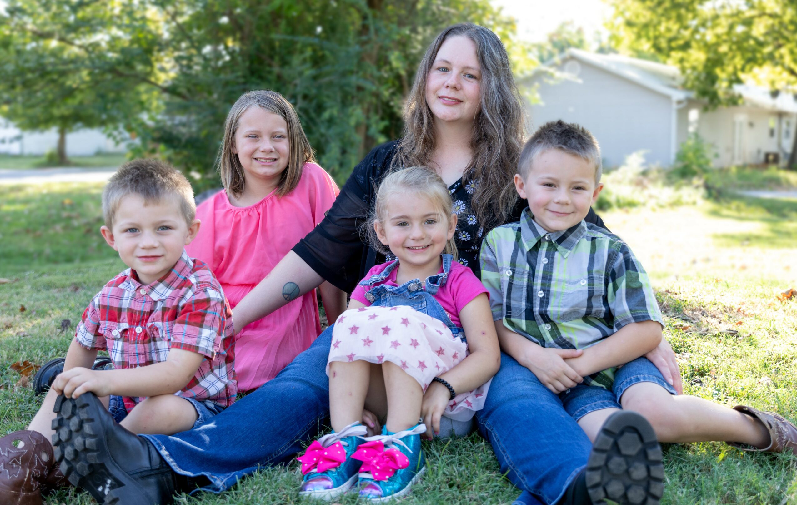 Group of five people, including a woman and four children, sitting outdoors on grass with trees and a house in background.