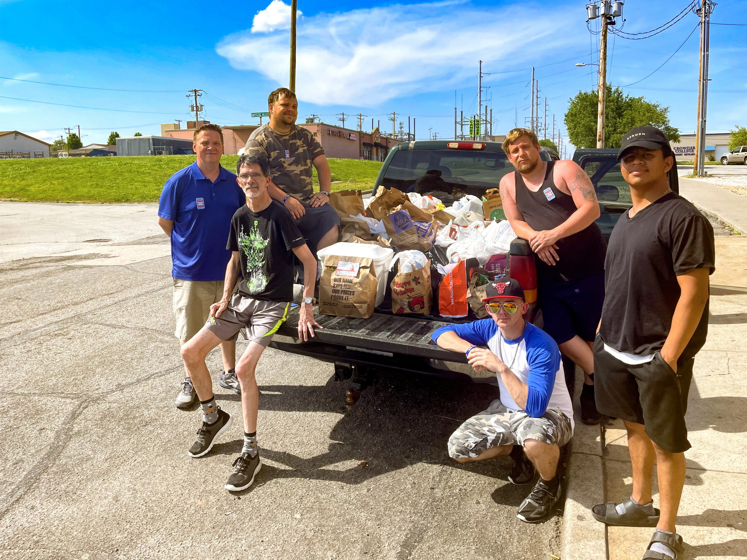 Group of six people standing and sitting near a pickup truck loaded with bags and supplies outdoors on a sunny day.