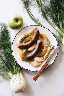 Plate with cooked sausages and onions, green apple, fennel bulb, and fresh herbs on white surface.