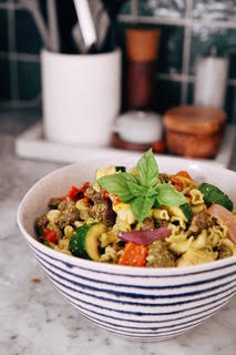Bowl of pasta salad with vegetables and basil garnish on a kitchen countertop, with utensils and containers in the background.