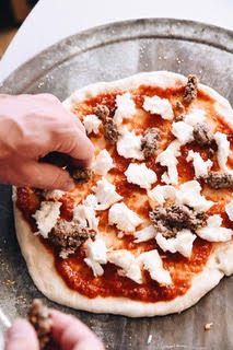Person adding cheese to a pizza with tomato sauce on a baking tray.