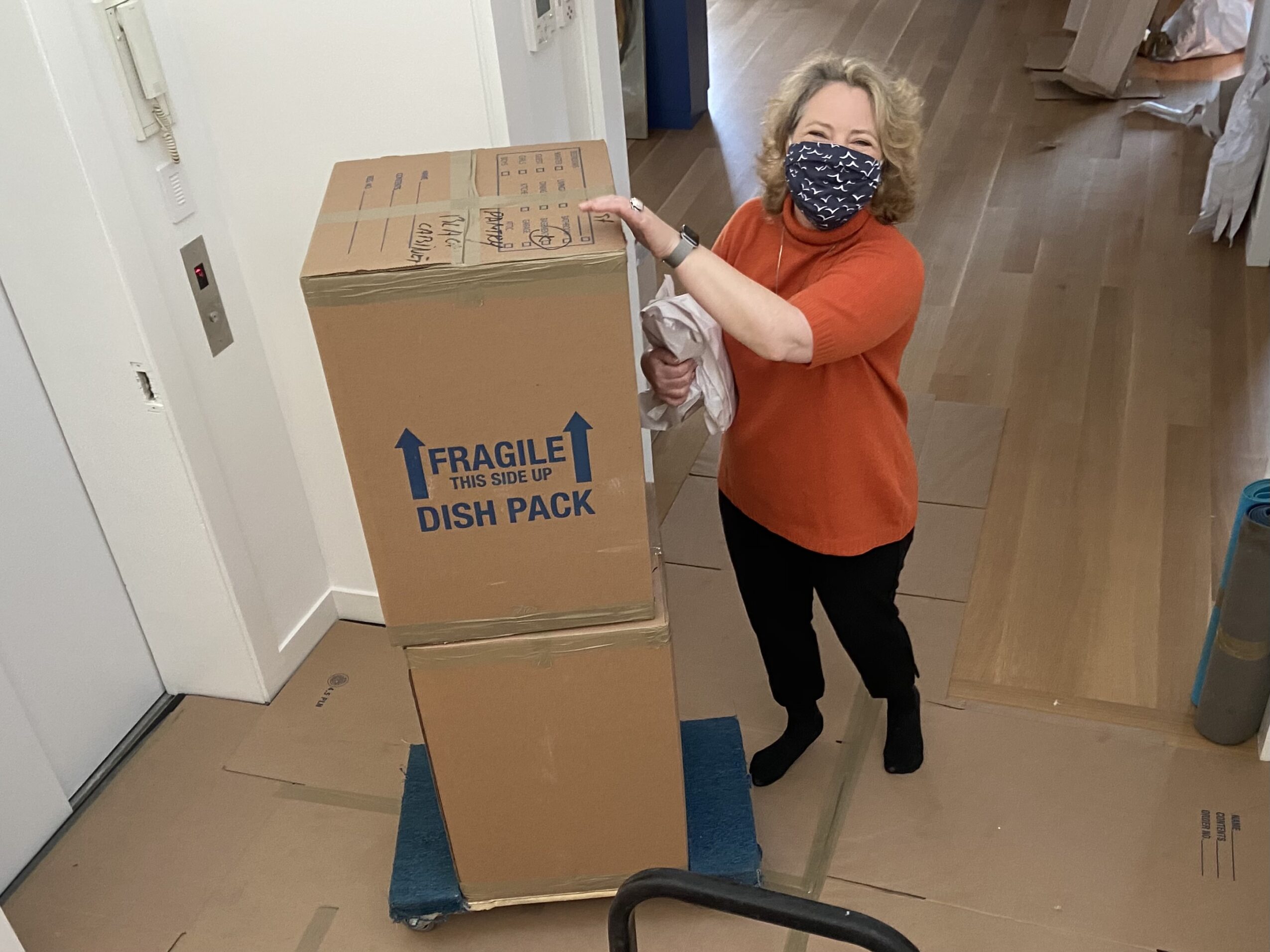 Woman wearing a face mask and orange top pushes a large cardboard box on a blue dolly in a room with wooden flooring.