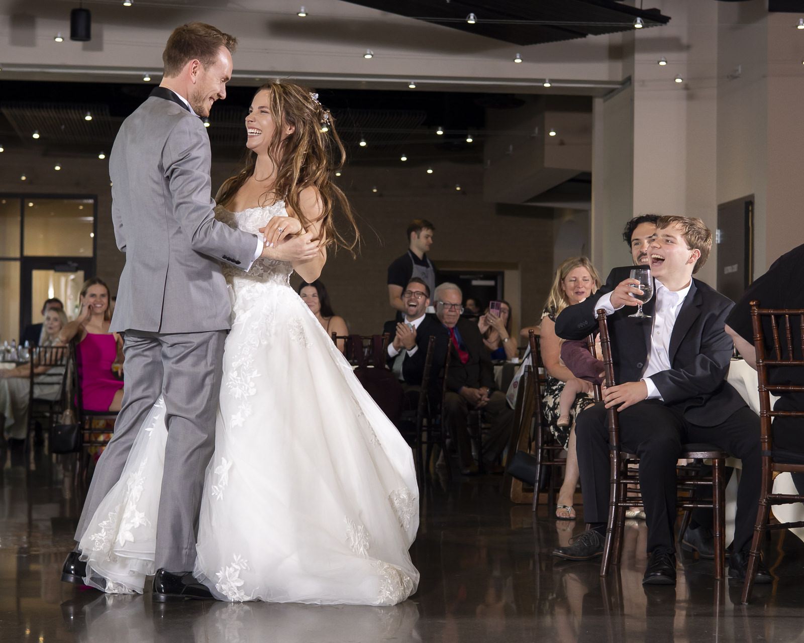 A bride and groom dance at their wedding reception, with guests seated at tables in the background.