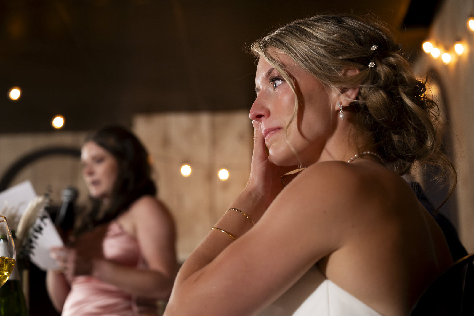 A woman with blonde hair in an updo, wearing a strapless dress, resting her face on her hand at an event.