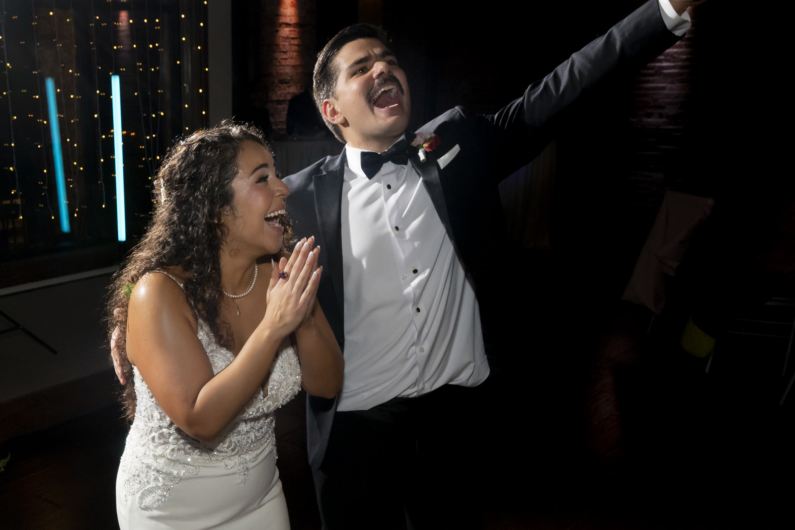 A man and woman in formal attire celebrating, with the man raising his arm and the woman clapping, in a dark setting.