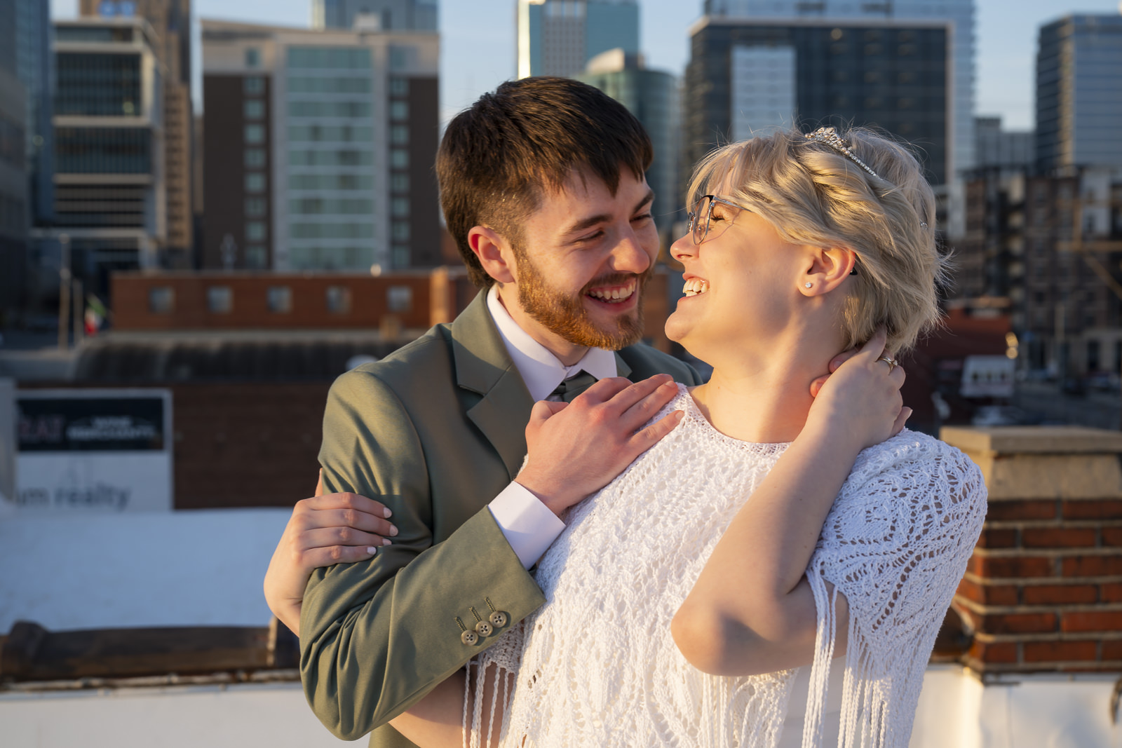 A smiling man and woman embrace outdoors with city buildings in the background.