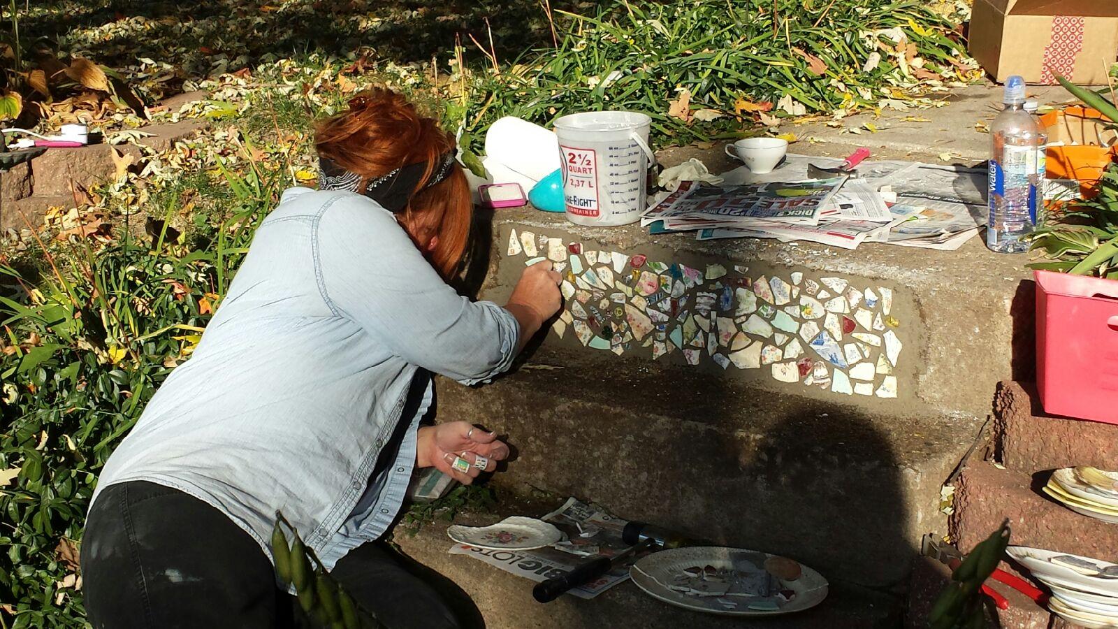 Person crouching outdoors, working on a mosaic table with various supplies nearby, surrounded by greenery.