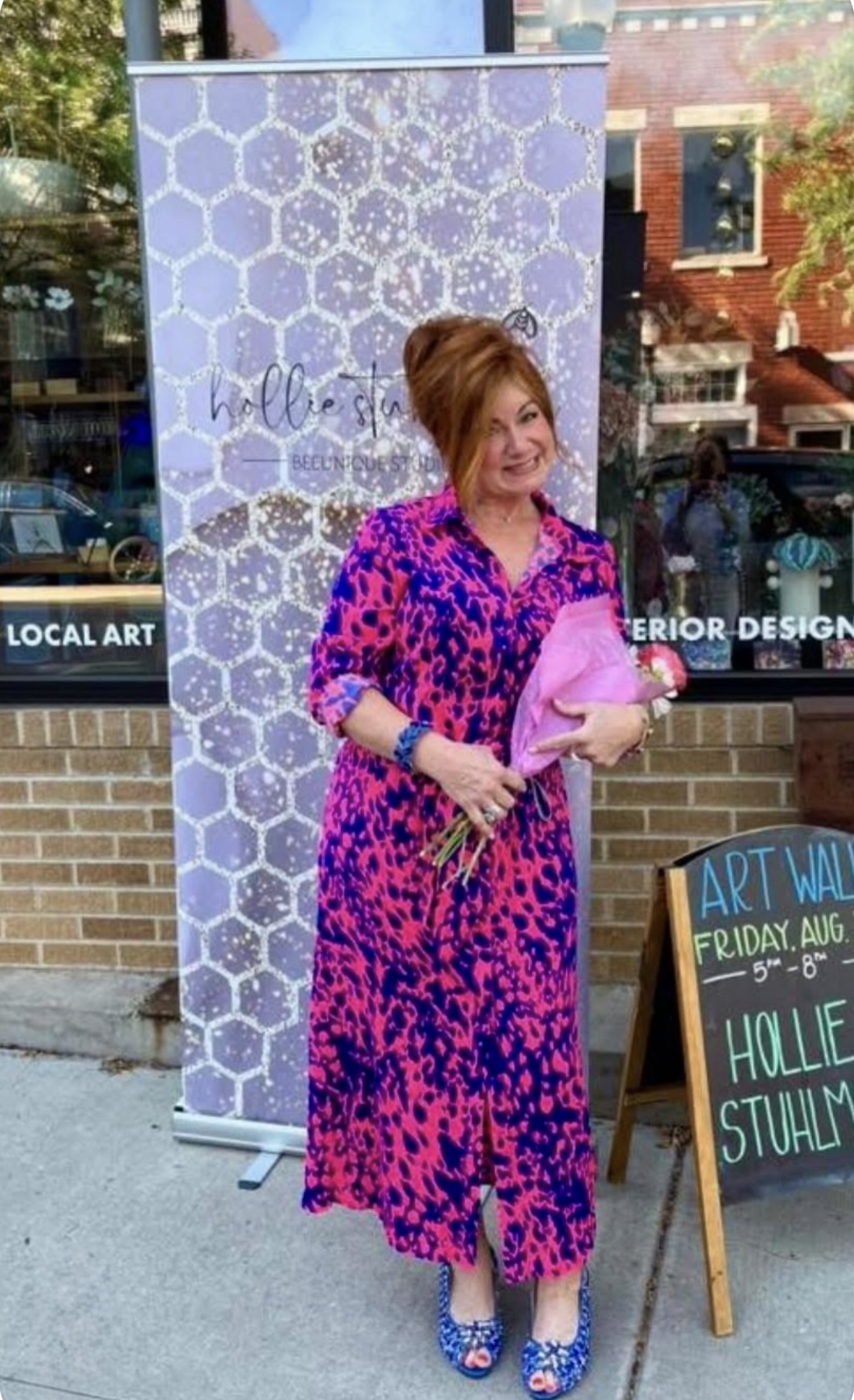 Woman in a pink and blue patterned dress holding a pink clutch, standing outside near a sign and art display.
