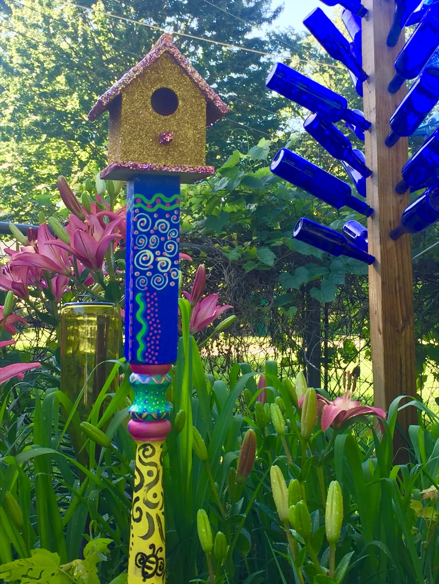 Colorful birdhouse on a decorated pole in a garden with pink flowers and green foliage.
