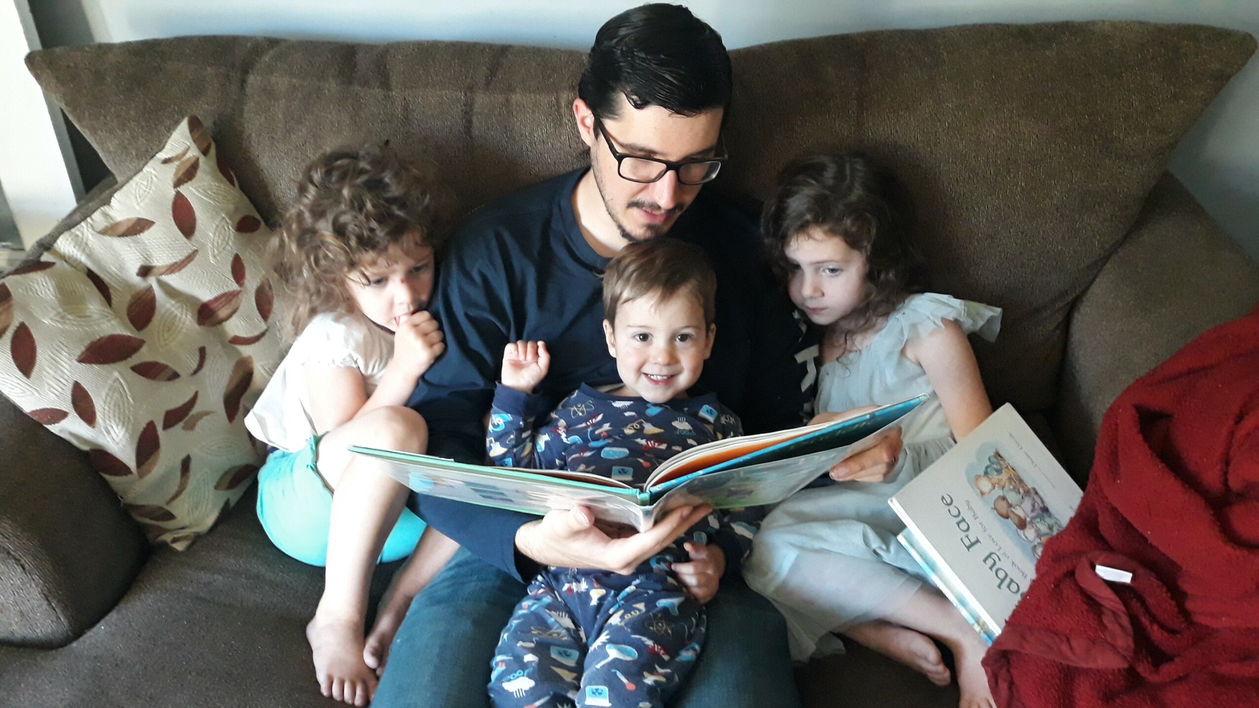 Man reading a book to three children on a sofa, with pillows around them.