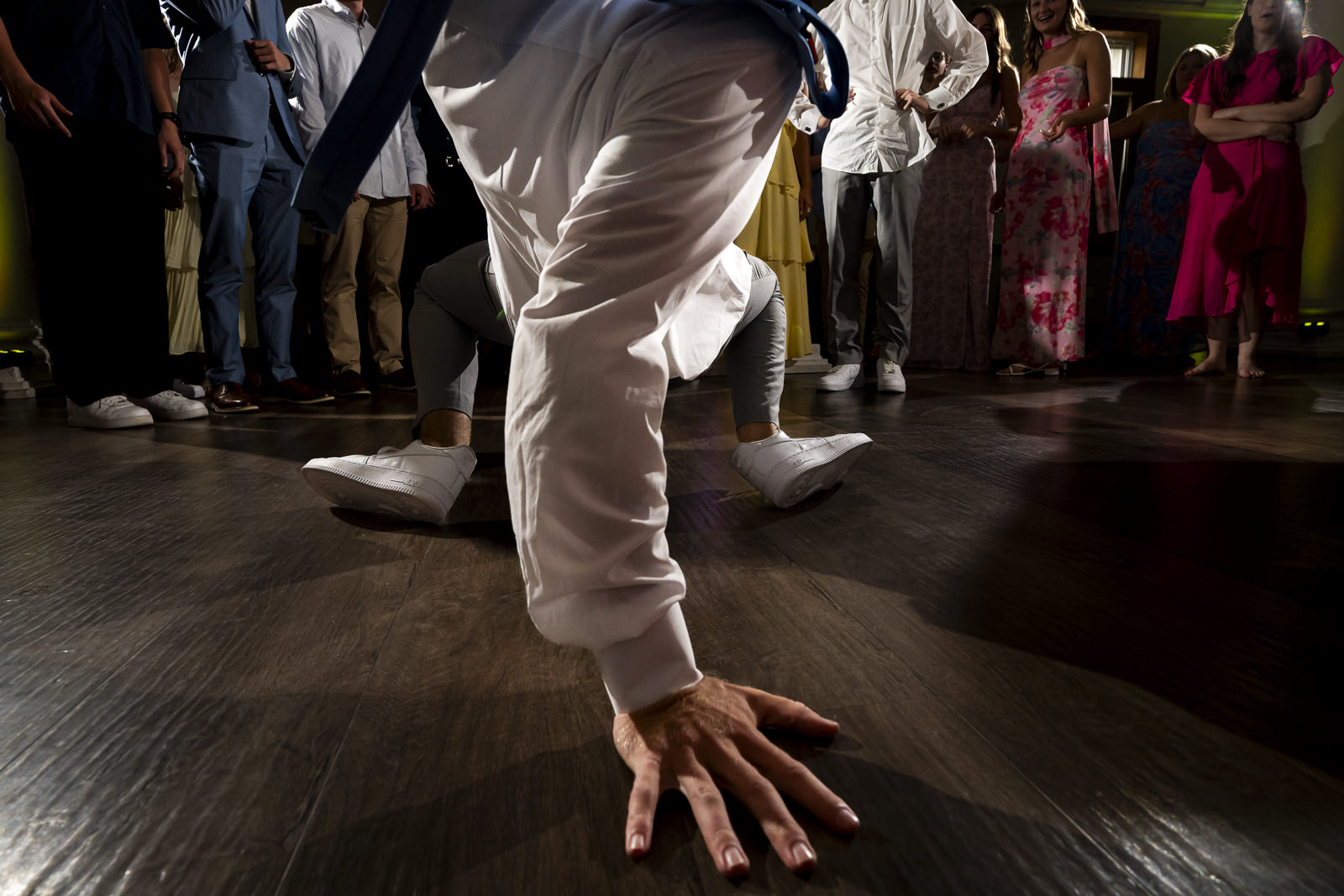 Person performing a handstand or breakdance move on a wooden floor with a group of people watching in the background.