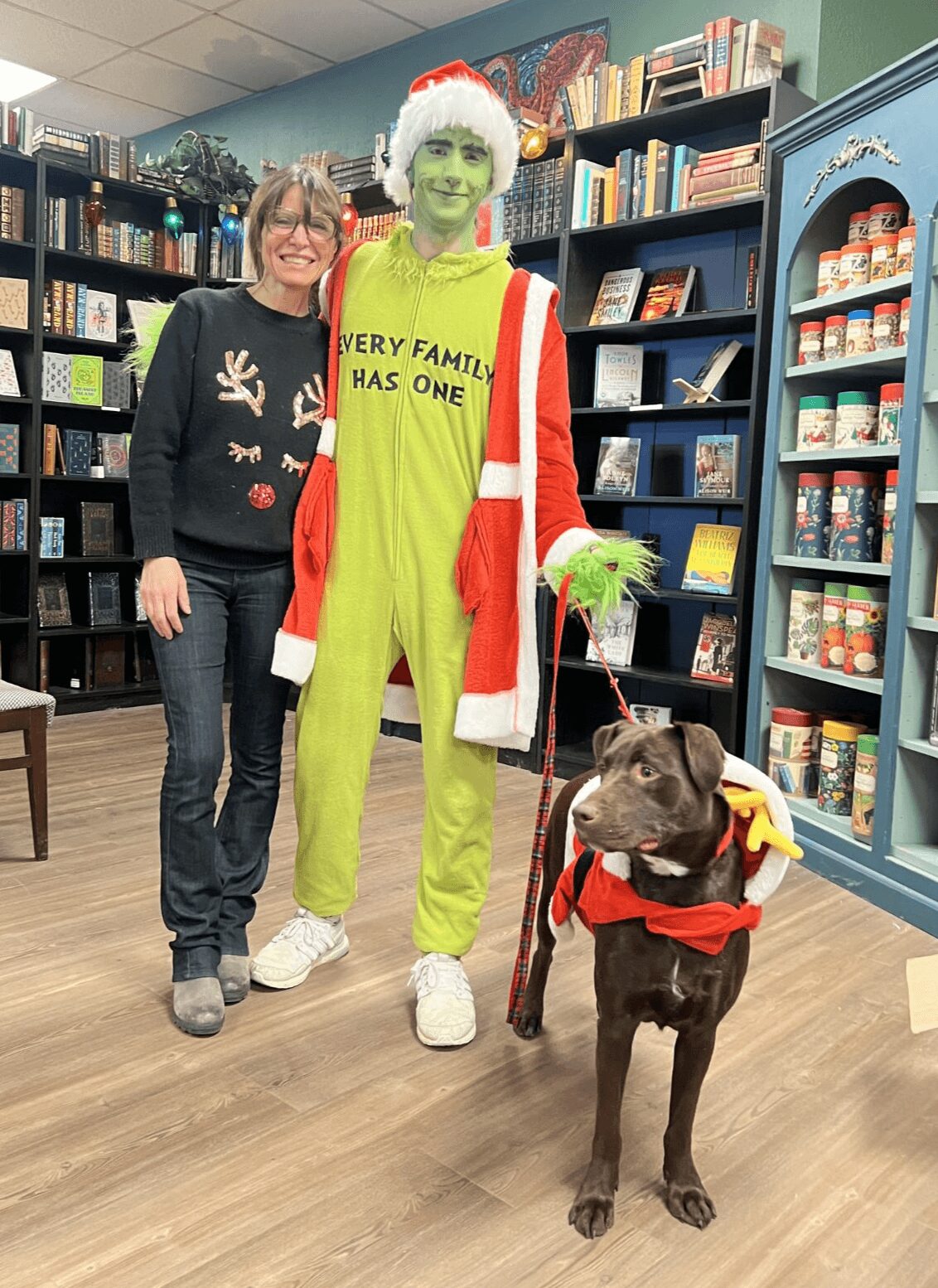 Two women and a person dressed as the Grinch with a dog in a store. The Grinch wears a Santa hat and red coat.