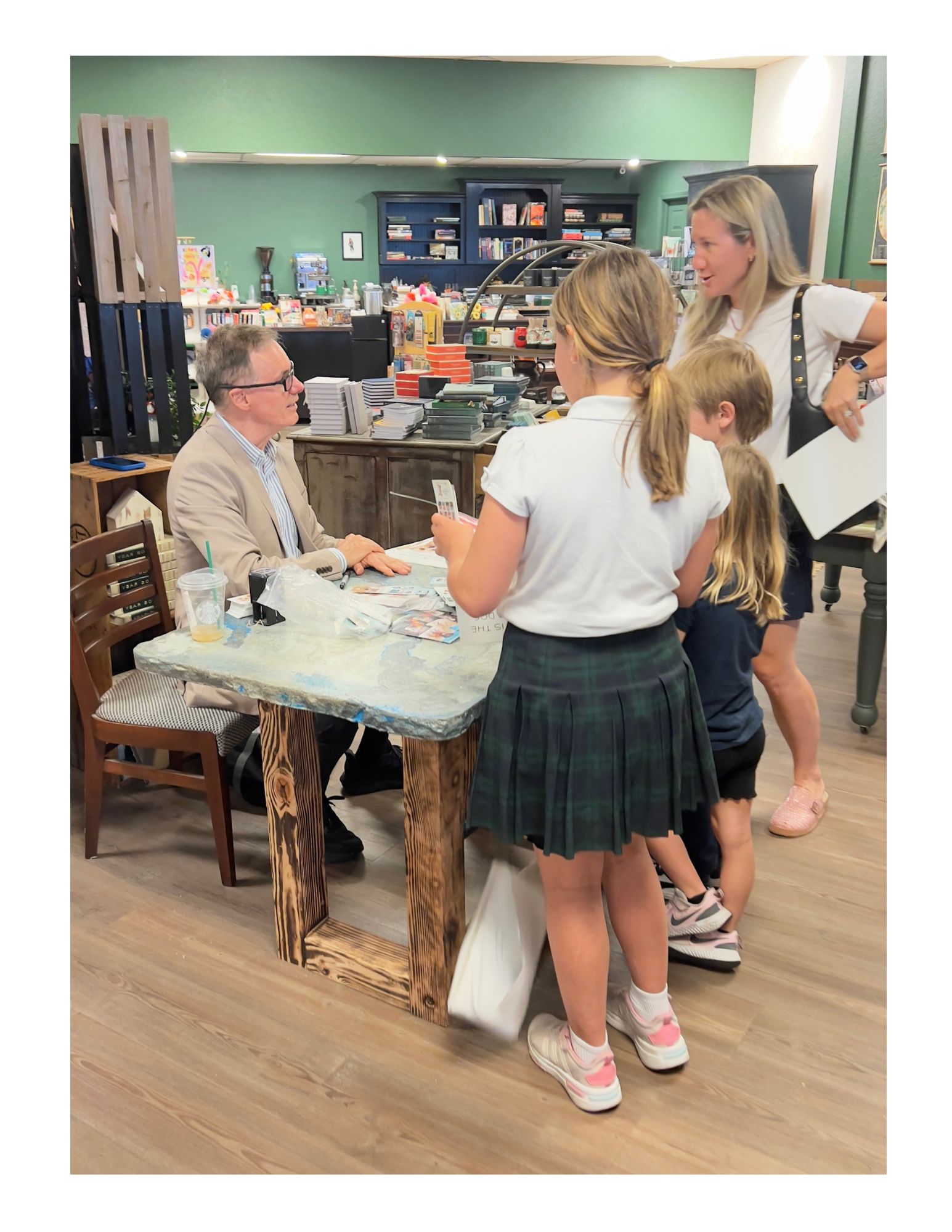 Children gather around a seated woman at a table in a store, with shelves and products in the background.