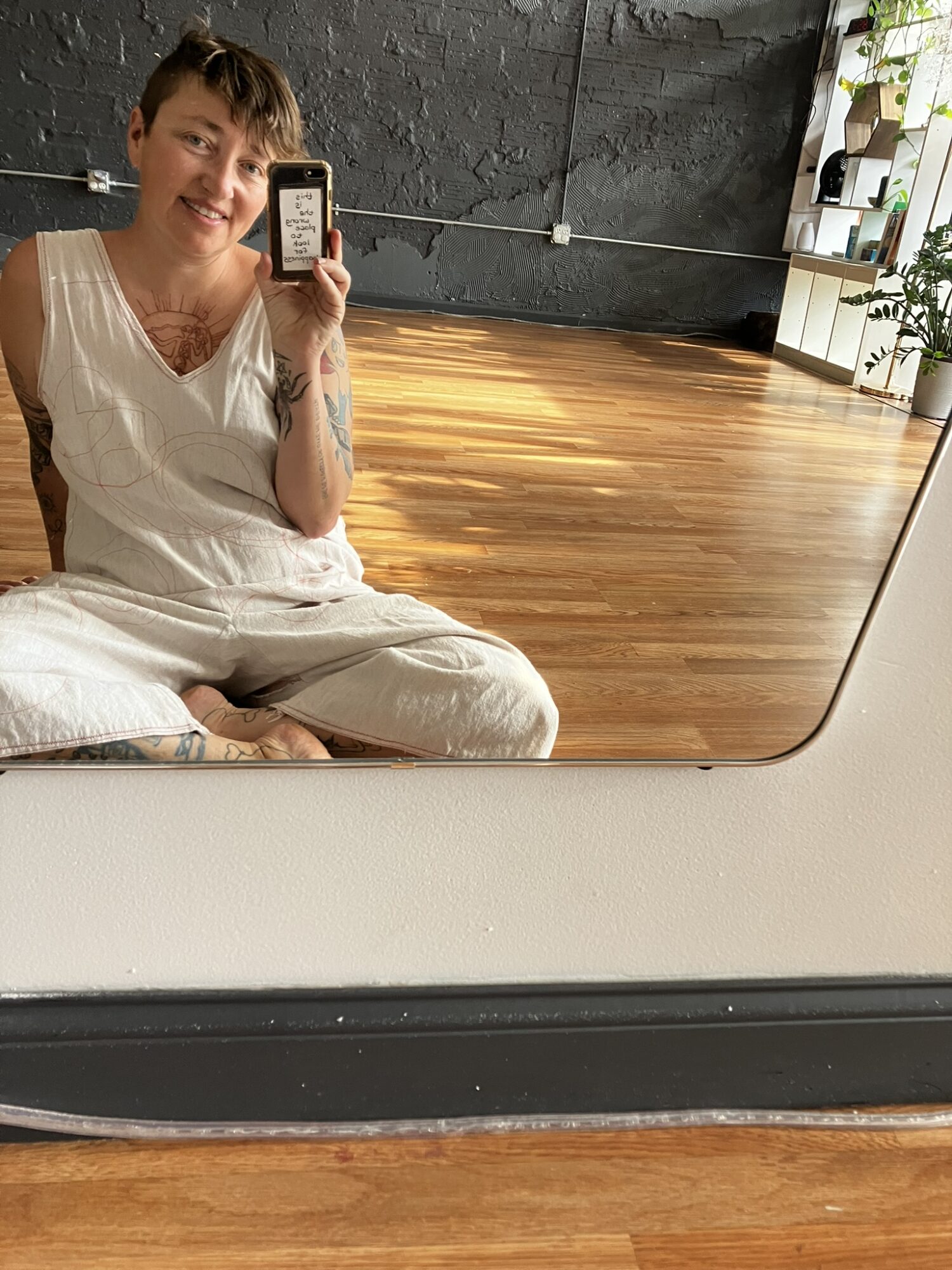 Person sitting on the floor taking a selfie in a mirror, with a wooden floor and industrial-style wall in the background.