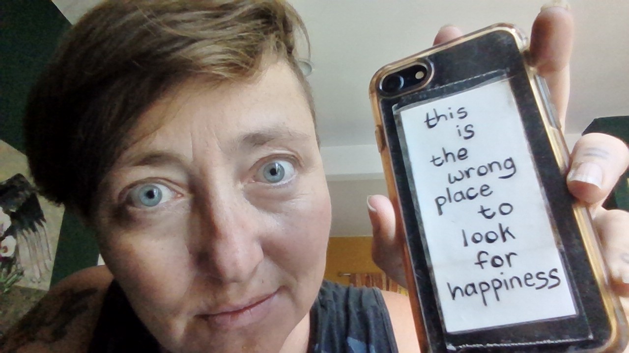 Person holding a phone with a handwritten note reading, 'this is the wrong place to look for happiness,' close-up face.