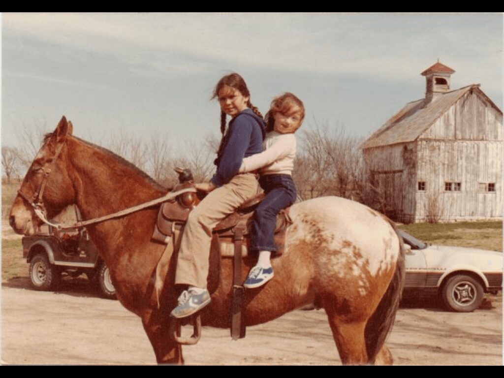 Two children sit on a horse, with a barn and cars in the background, outdoors during daytime.