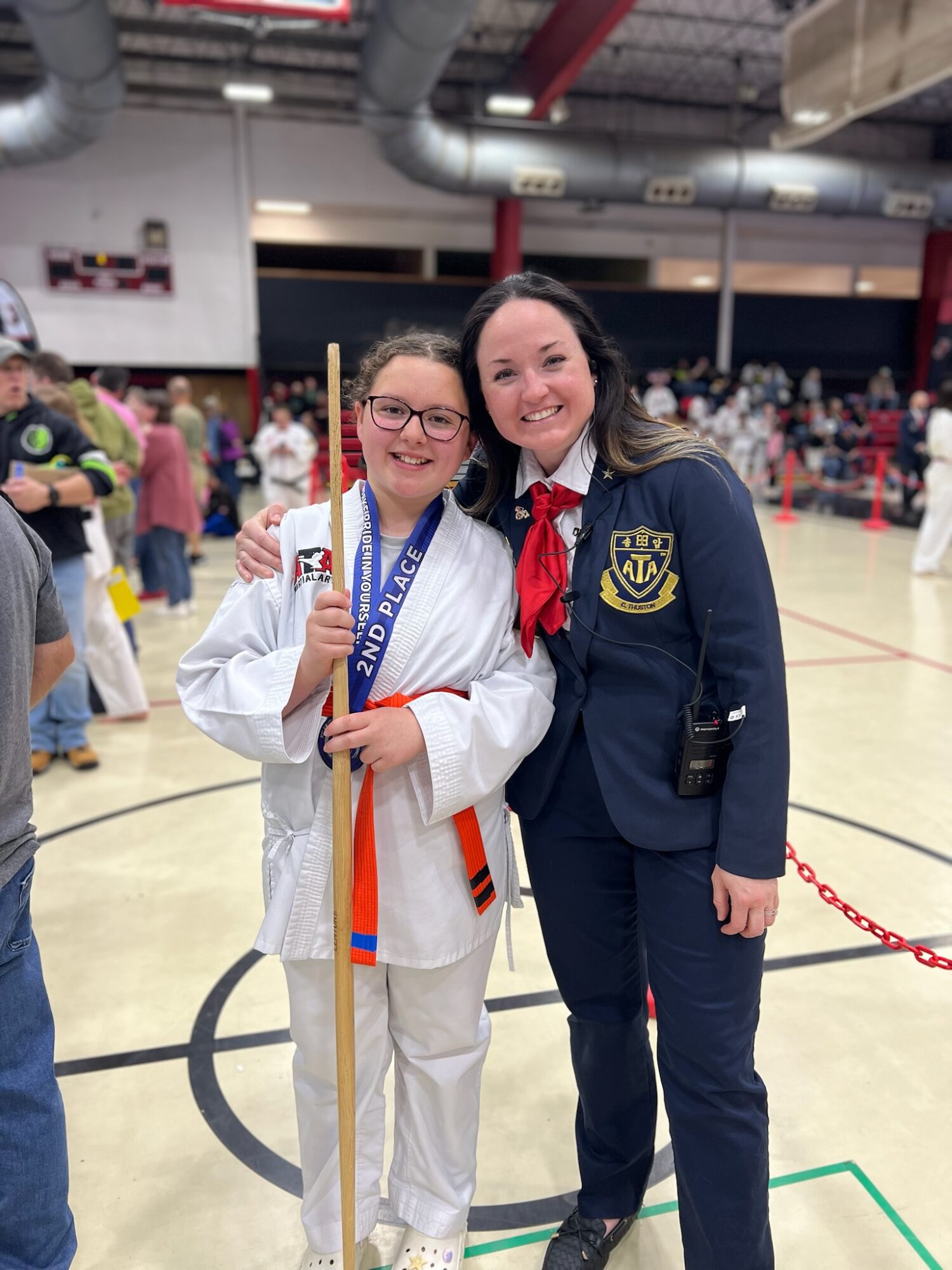 Two women, one in a white martial arts uniform holding a medal and a stick, and another in a dark blazer, smiling together in a gymnasium.