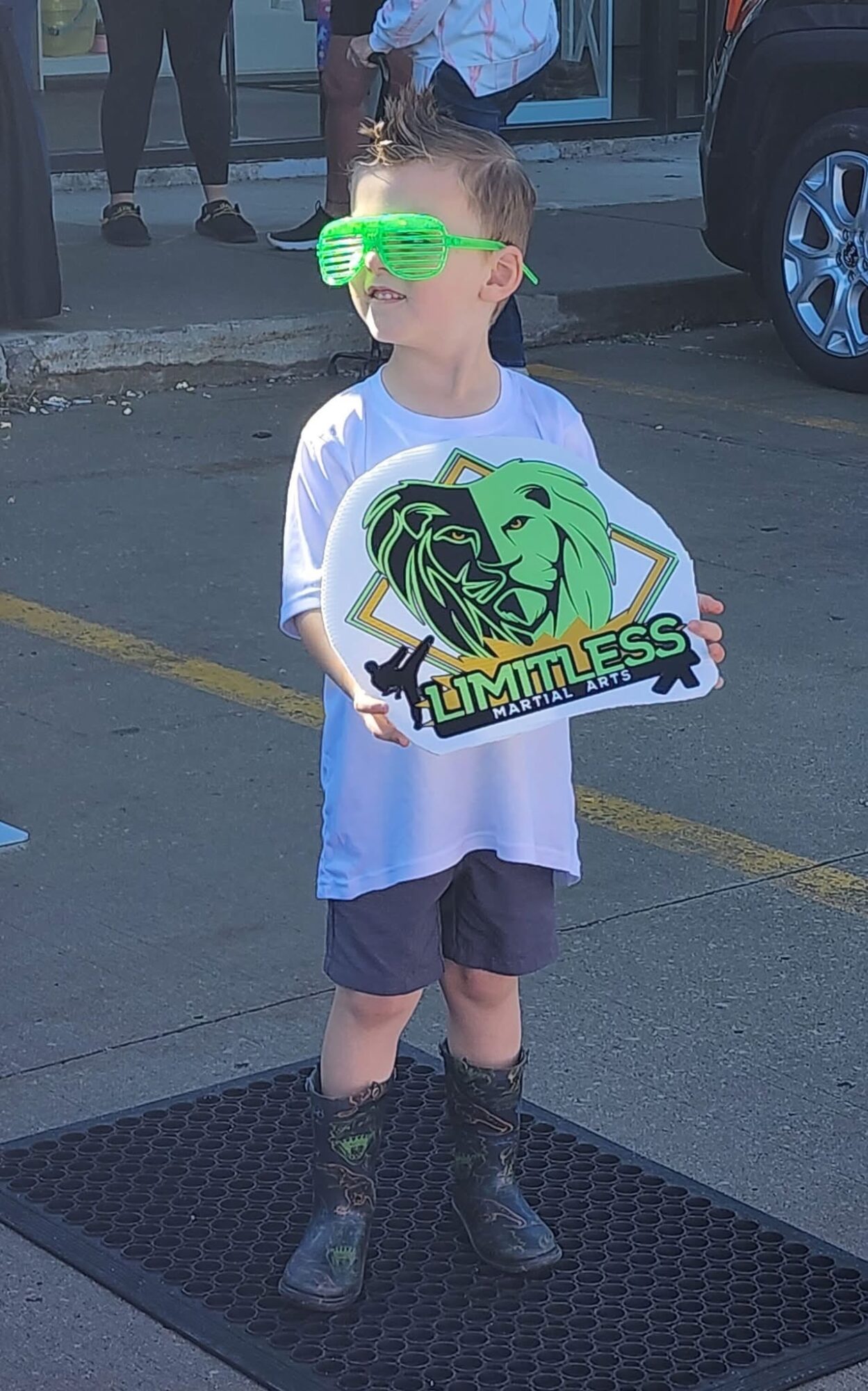 Child wearing green sunglasses holding a sign with a lion and the word 'Limitless' in a parking lot.