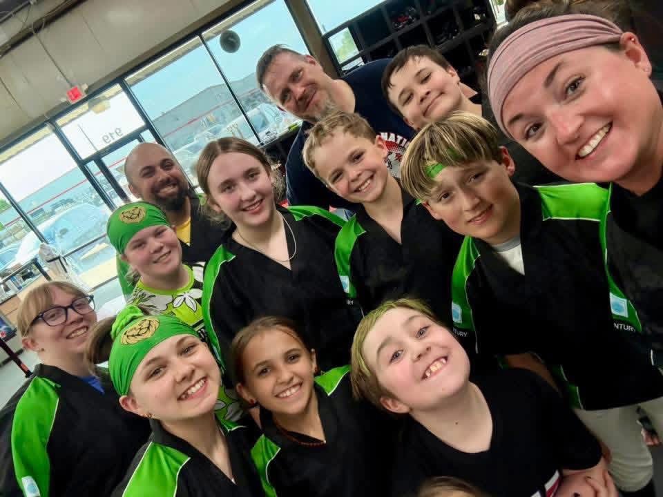 Group of children and adults smiling indoors, some wearing green headbands and black jackets with green accents.