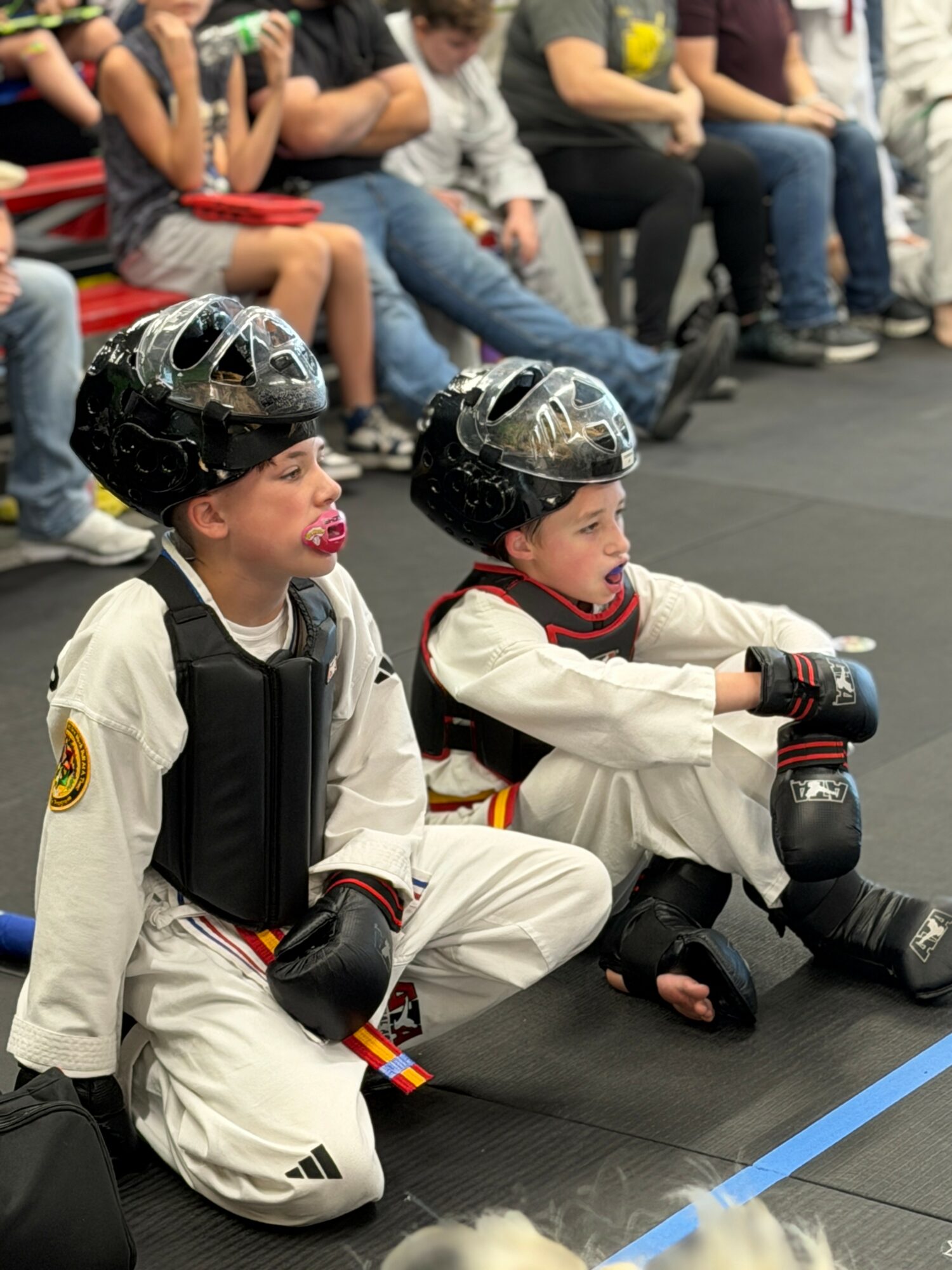 Two children in martial arts uniforms and protective gear sit on the floor, watching an event, with a group of people seated behind them.
