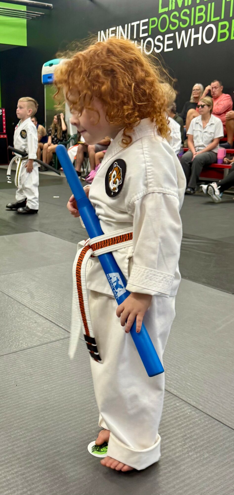 Child with curly hair in martial arts uniform holding a blue foam stick, standing on a mat with people in background.
