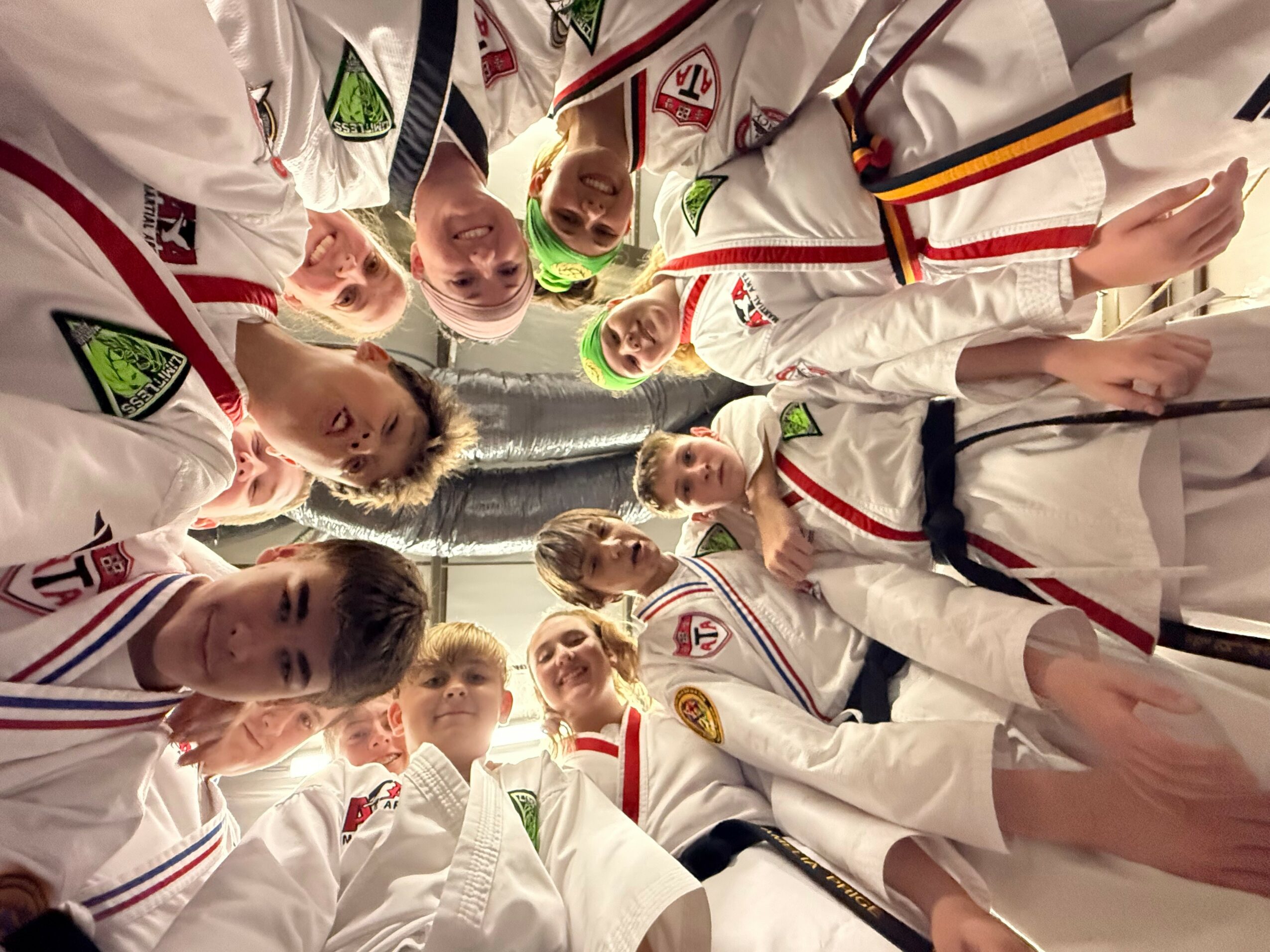 Group of children in martial arts uniforms standing in a circle, looking down at the camera, smiling.