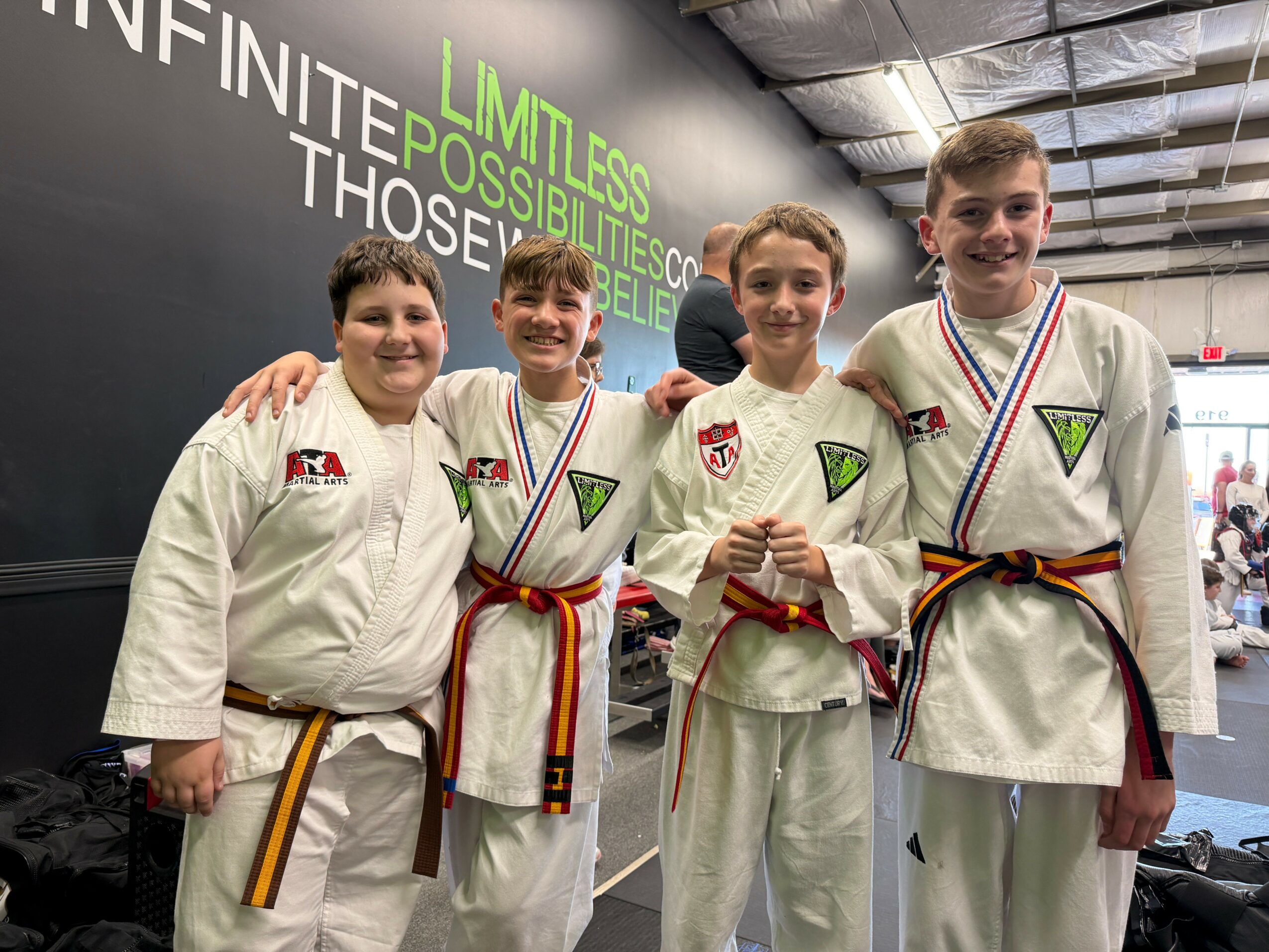Four young people in martial arts uniforms with medals, standing together indoors, smiling, with a black wall and motivational text behind them.