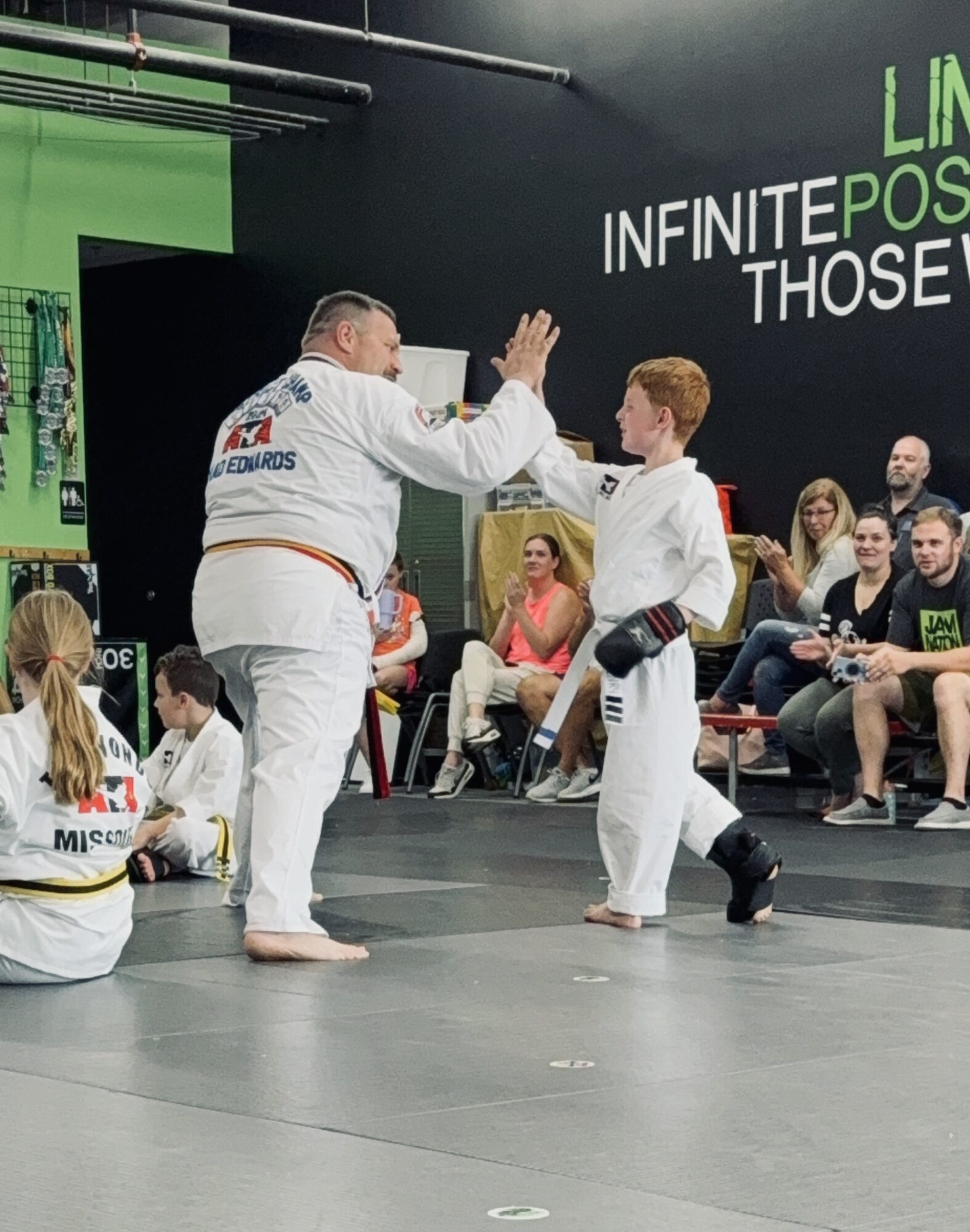 Two people in martial arts uniforms high-five in a gym with spectators seated around the mat.