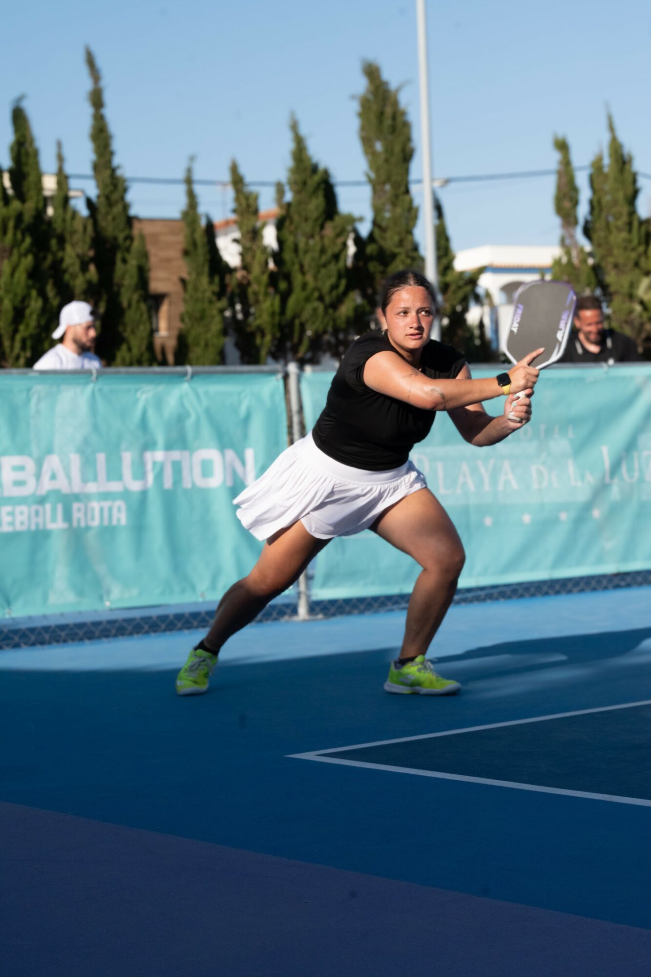 Female tennis player in black shirt and white skirt prepares to hit a ball on blue court outdoors.